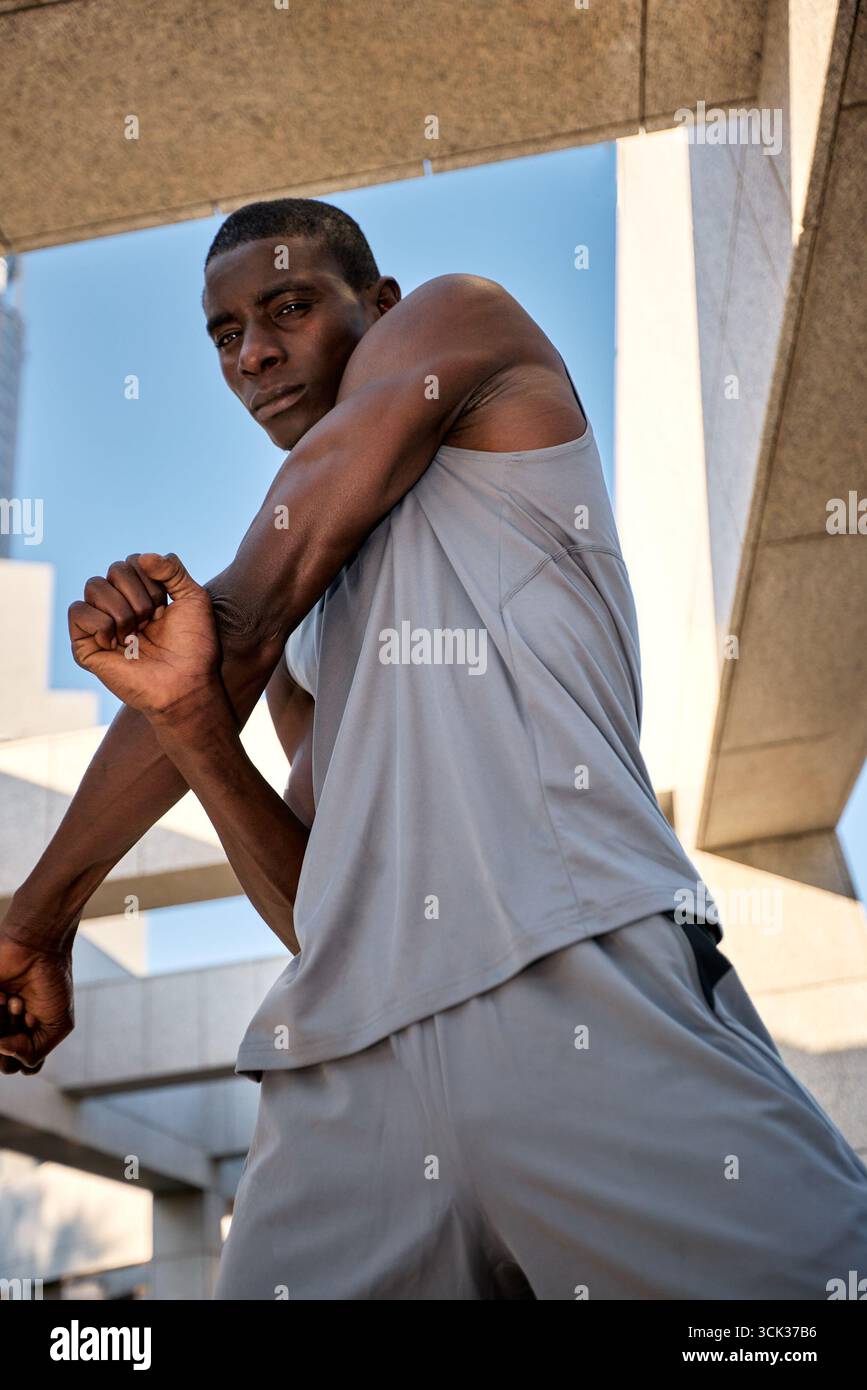 Un giovane in abbigliamento fitness viene visto mentre esegue un esercizio di stretching all'aperto. L'ambiente presenta strutture architettoniche urbane e un blu chiaro Foto Stock