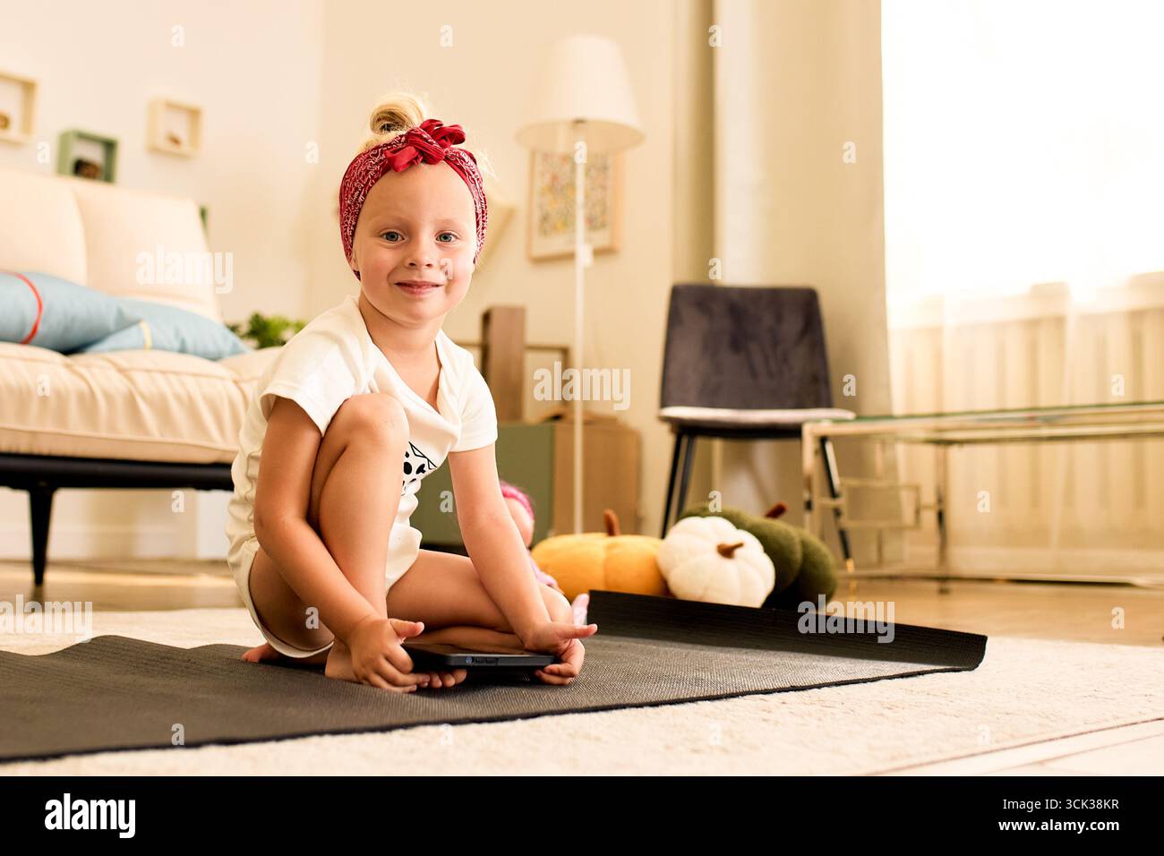 Una giovane ragazza allegra siede con fiducia su un tappetino da yoga in un soggiorno splendidamente illuminato. Con un sorriso cordiale, l'arredamento accogliente la circonda, creando una guerra Foto Stock