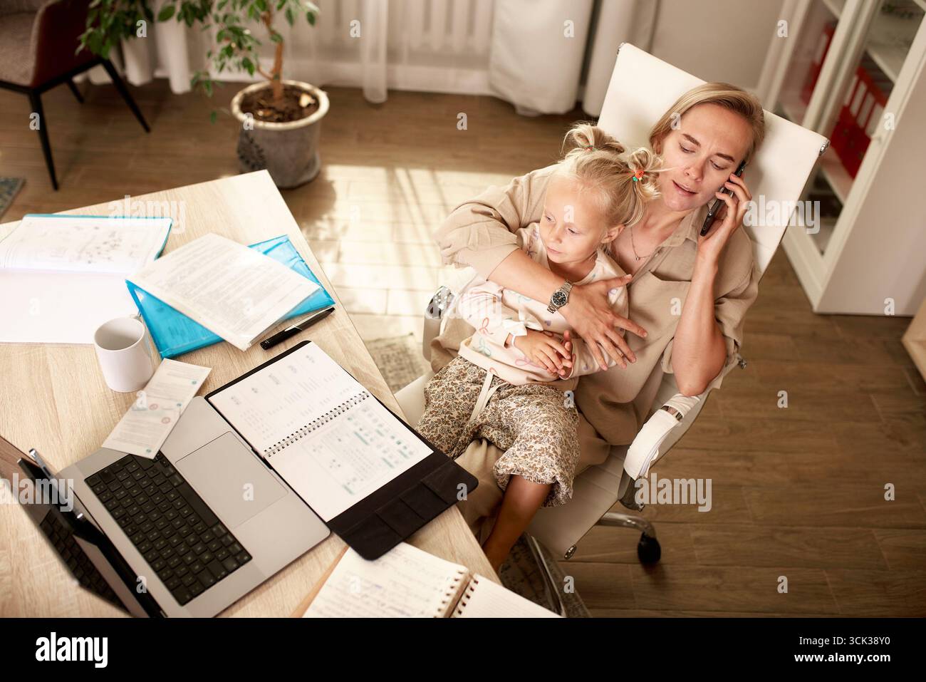 Una madre lavora in remoto dal suo ufficio domestico, multitasking tra genitori e lavoro. È seduta a una scrivania e gestisce una conversazione al telefono W Foto Stock