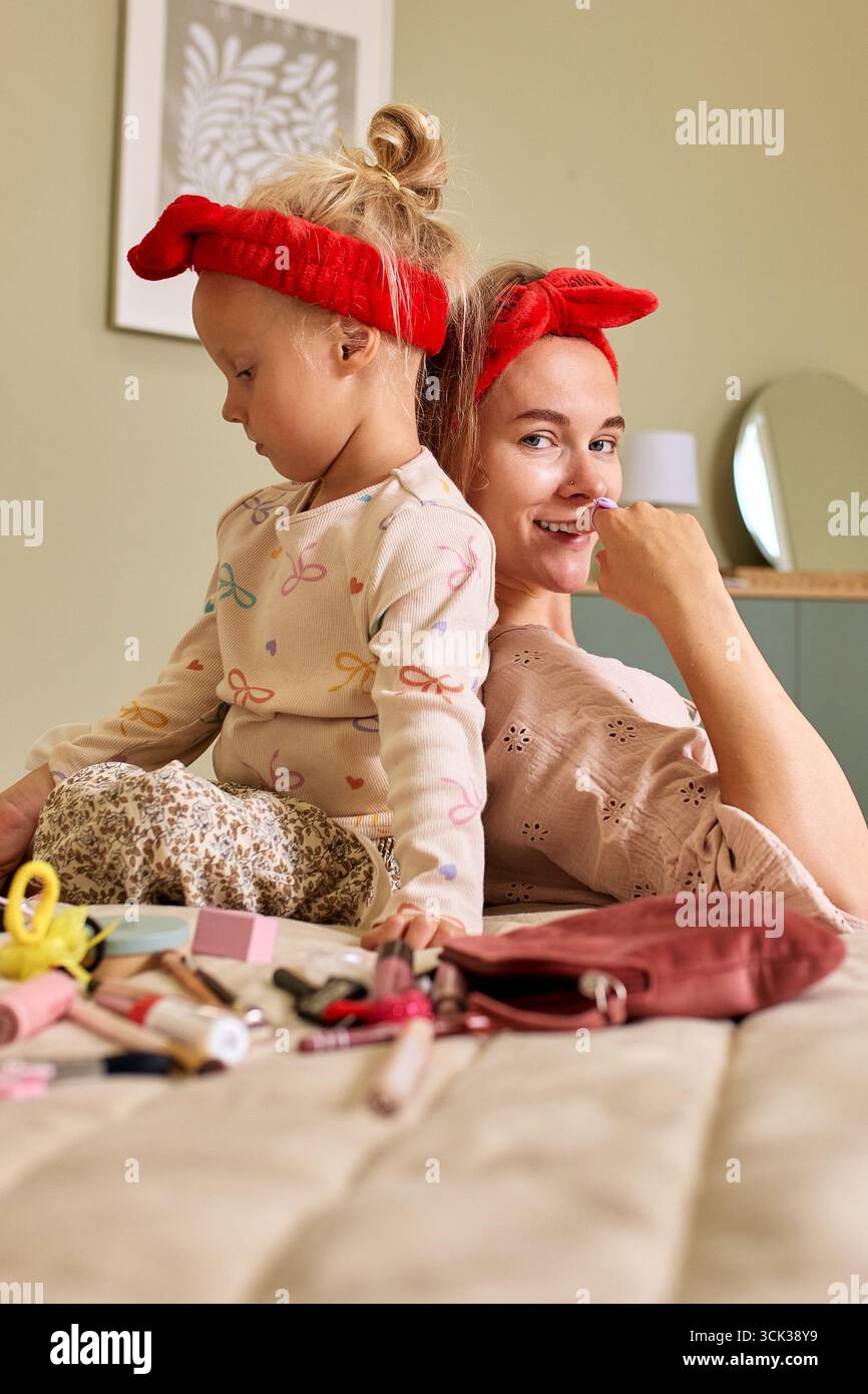 Una madre allegra e sua figlia trascorrono del tempo insieme in una camera da letto ben illuminata, esplorando il trucco e divertendosi. Entrambi i modelli di usura h Foto Stock