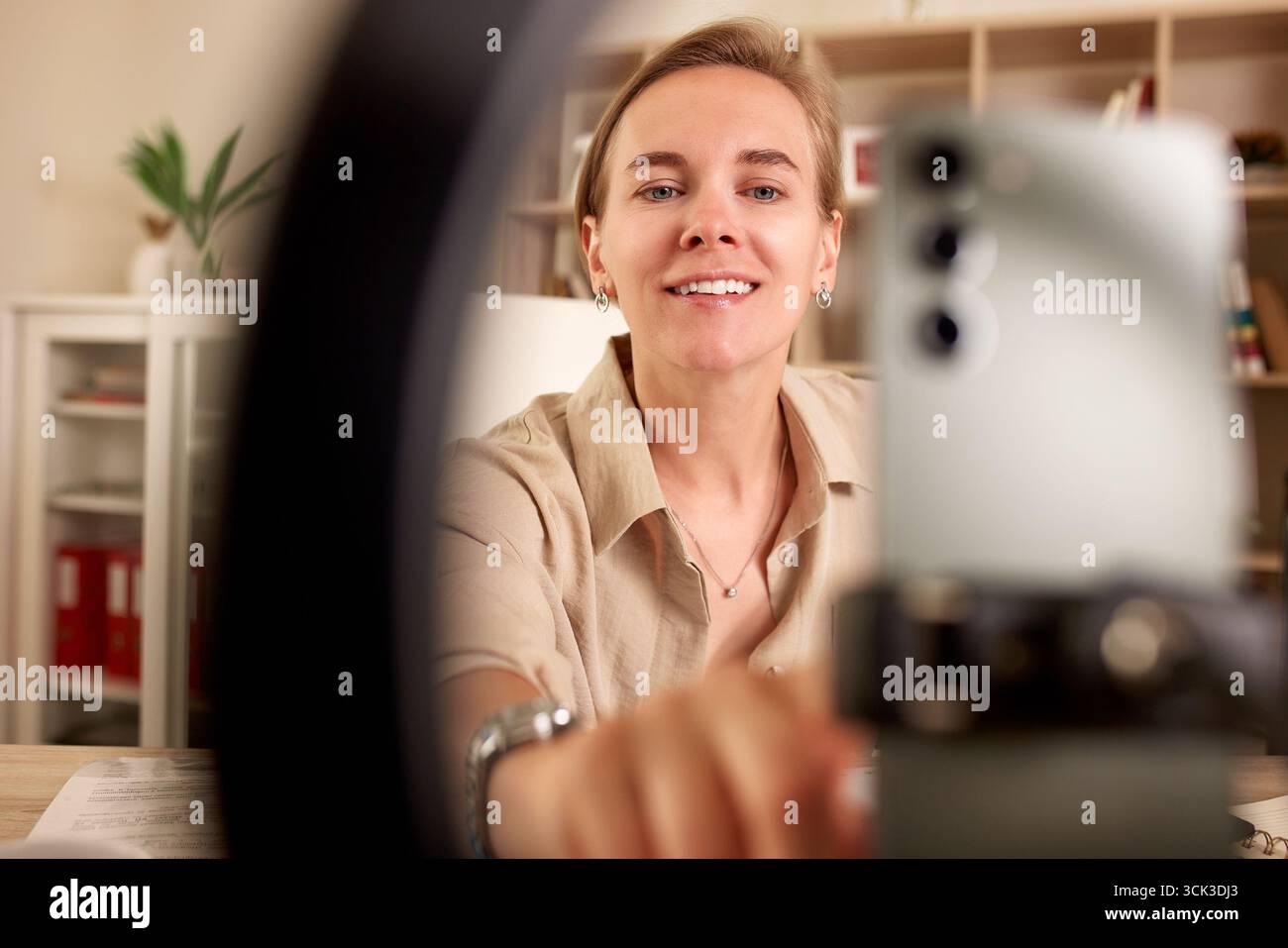 Una donna allegra interagisce con il suo telefono per una sessione video in un piacevole spazio interno. La sua espressione gioiosa e il suo abbigliamento casual creano un'invitante Foto Stock