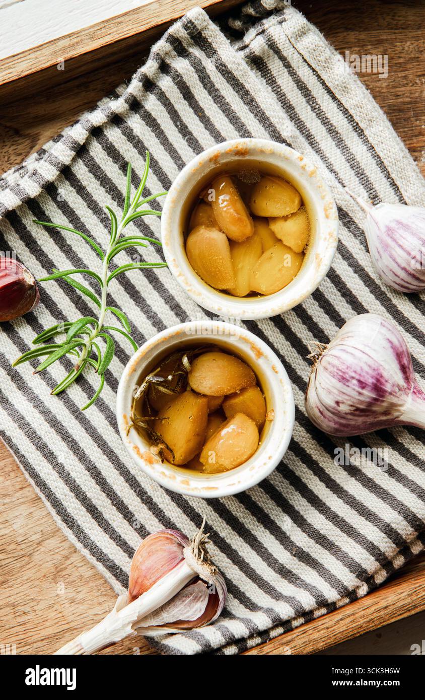 Vista dall'alto del confit di aglio arrosto del forno all'interno di piccole pentole da forno su un asciugamano da cucina. Guanti in rami di rosmarino e aglio per la decorazione. Foto Stock