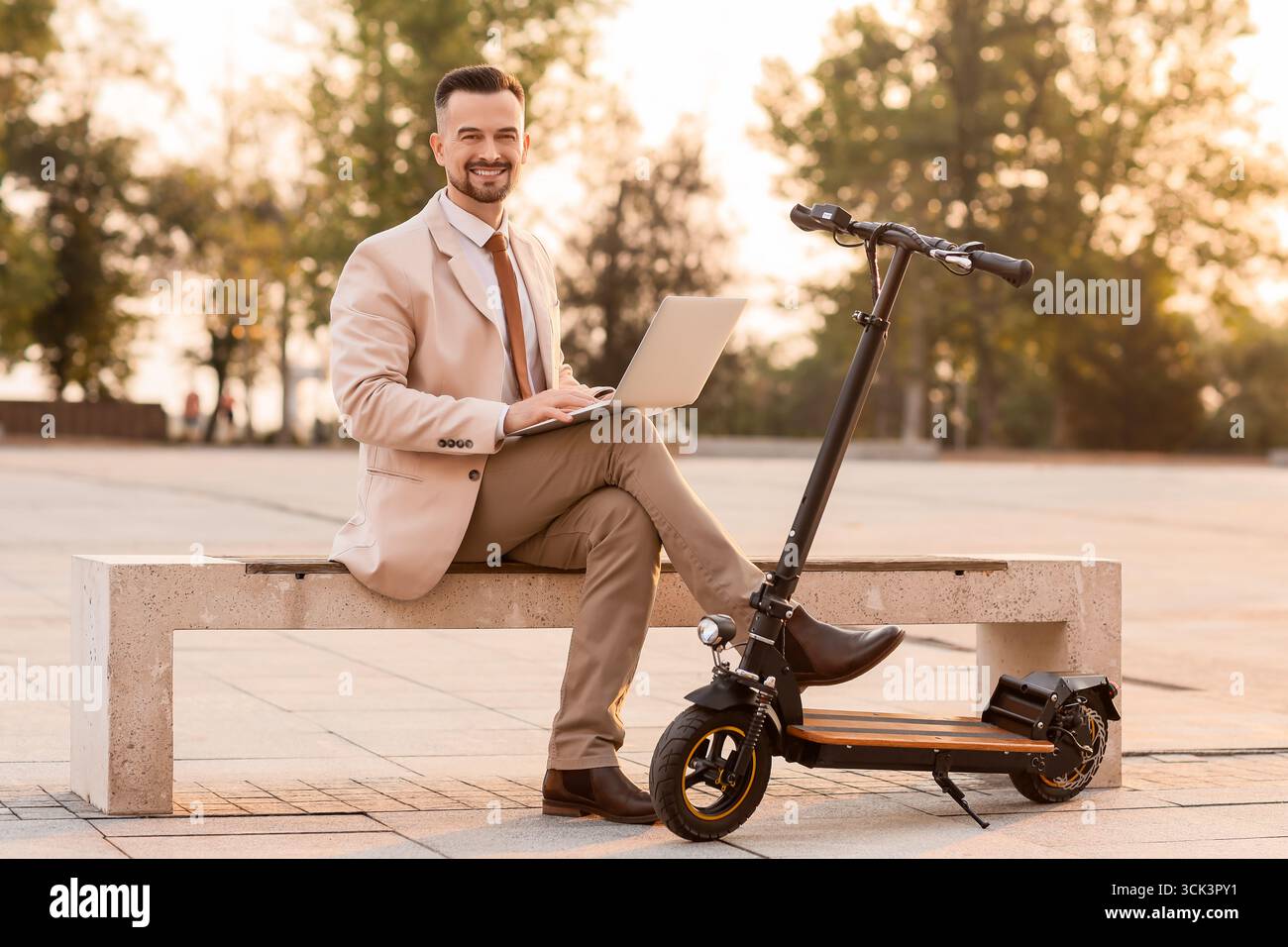 Bellissimo giovane uomo d'affari con scooter elettrico e laptop seduto sulla panchina nel parco Foto Stock
