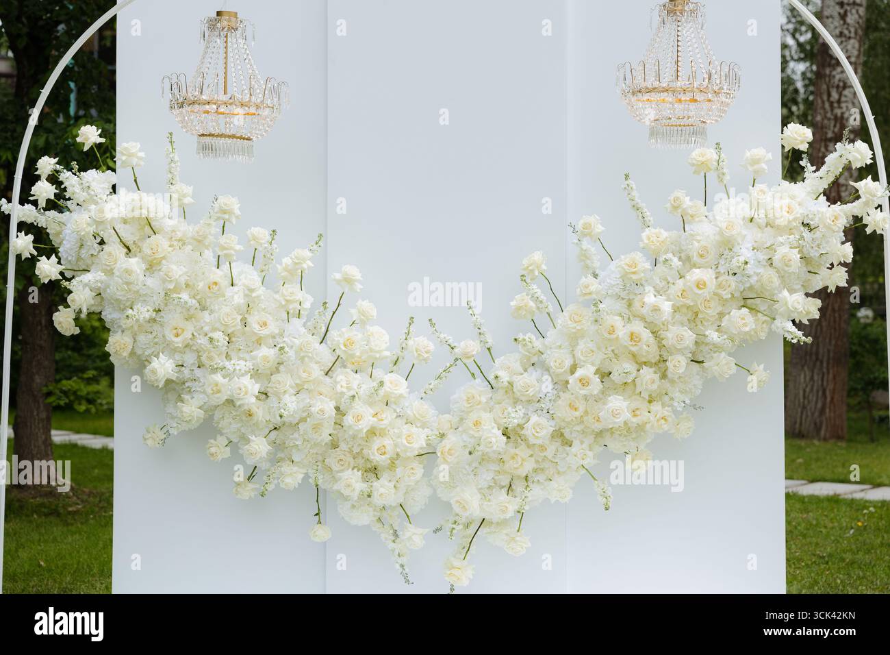 Uno splendido spettacolo floreale di rose bianche e delicati fiori decora uno sfondo verde, impreziosito da lampadari sospesi, evocando eleganza. Foto Stock