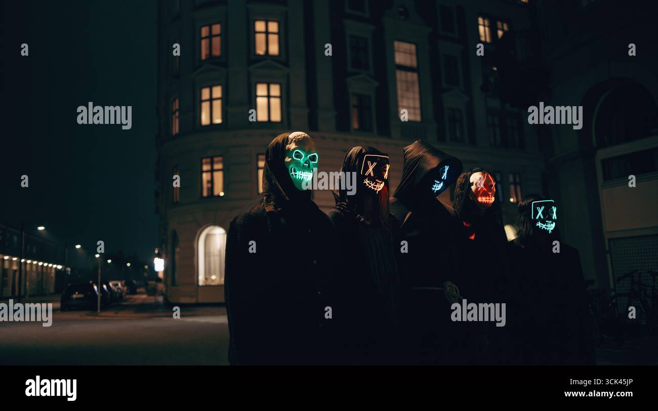 Un gruppo di amici vestiti da personaggi inquietanti con maschere al neon brillanti, che si riuniscono in una strada urbana poco illuminata di notte per festeggiare Halloween in un moo Foto Stock