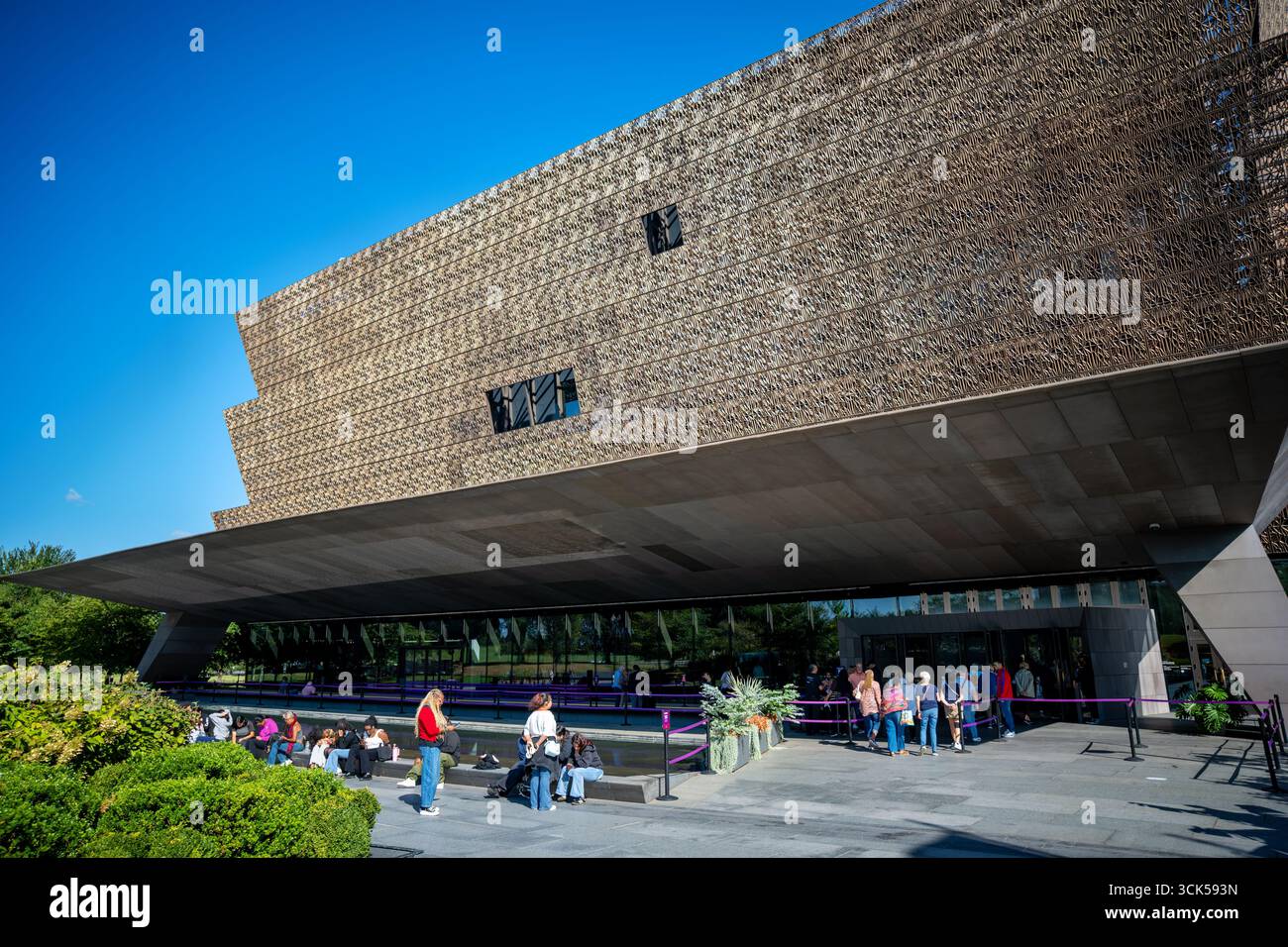 National Museum of African American History and Culture Washington DC // WASHINGTON DC - il National Museum of African American History and Culture è visibile in una giornata di sole. La caratteristica facciata color bronzo del museo presenta un complesso motivo geometrico. I visitatori vengono visti riuniti fuori dall'edificio, alcuni seduti su panchine e altri in fila. Il museo, parte dello Smithsonian Institution, si trova sul National Mall ed è stato inaugurato nel 2016. È dedicato a documentare la vita, la storia e la cultura afroamericana. Foto Stock