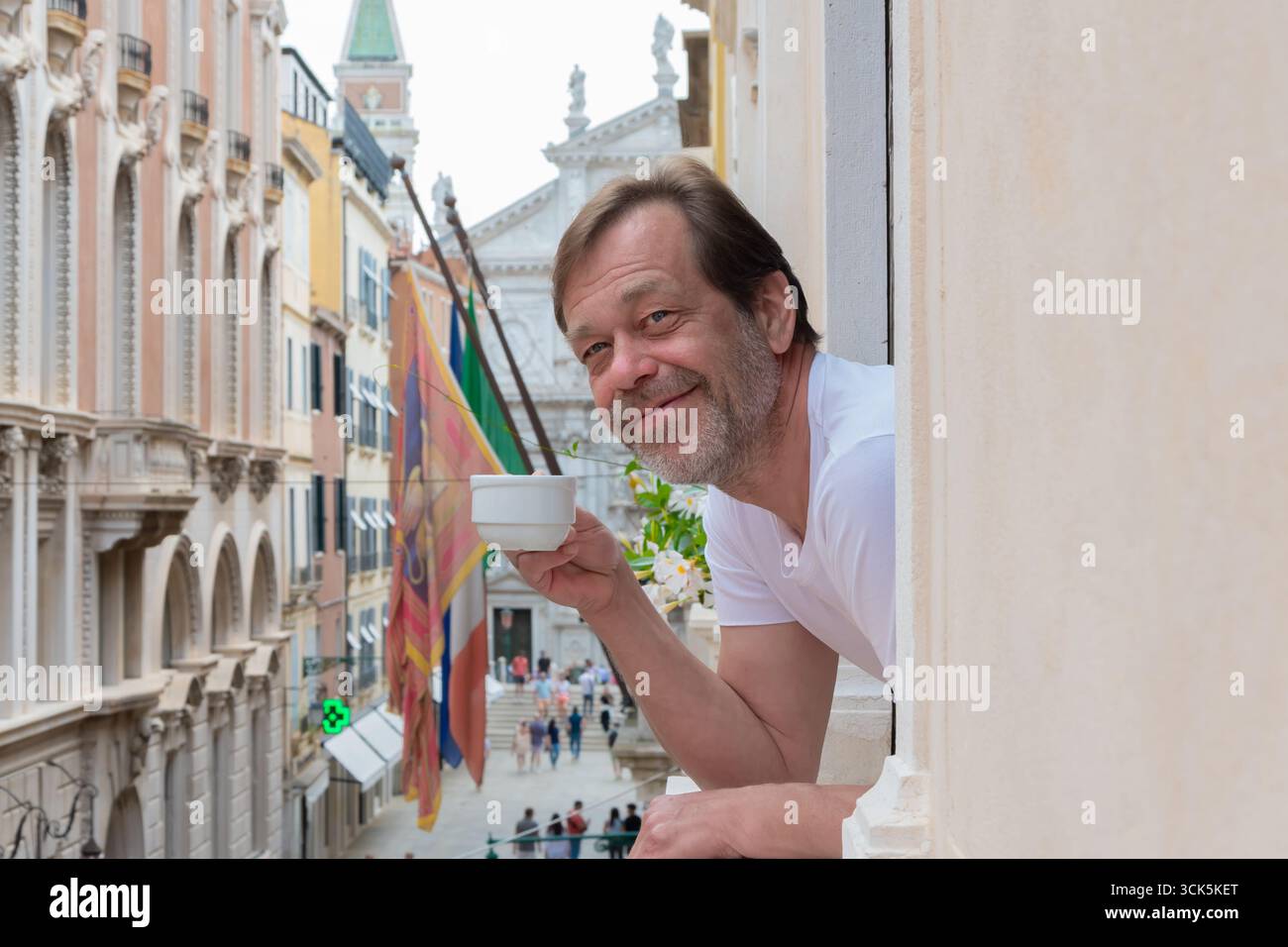 Un uomo con una tazza di caffè si trova alla finestra che guarda fuori dal balcone sulle strade del centro storico di Venezia Foto Stock