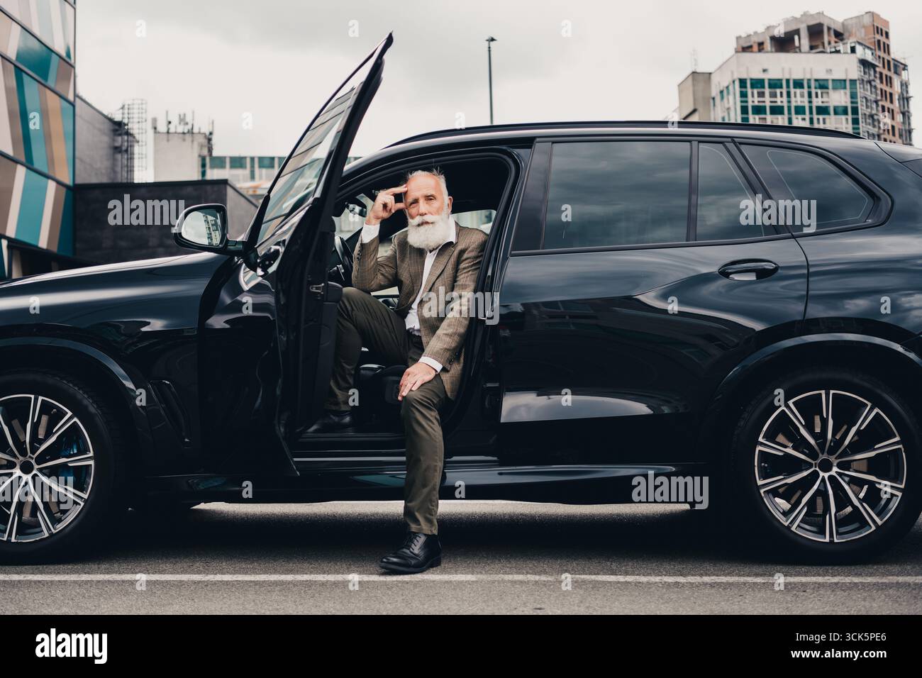 Elegante uomo d'affari anziano che posa sulle strade della città accanto a un'auto nera di lusso durante una giornata intensa Foto Stock