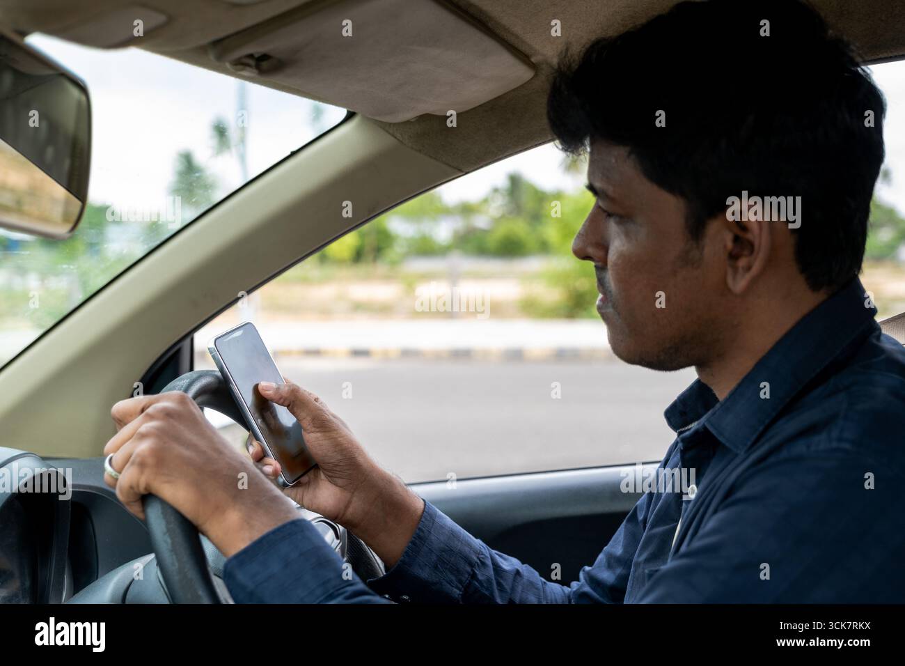 Il conducente controlla gli avvisi telefonici durante la guida su una strada trafficata, indicando una guida distratta in condizioni di sole. Foto Stock
