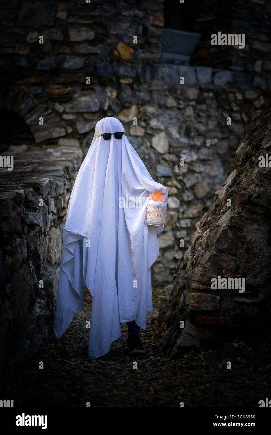 Un bambino in un foglio bianco, occhiali da sole e una lanterna in mano raffigura un fantasma sullo sfondo di un muro di pietra di un antico edificio. Halloween Foto Stock