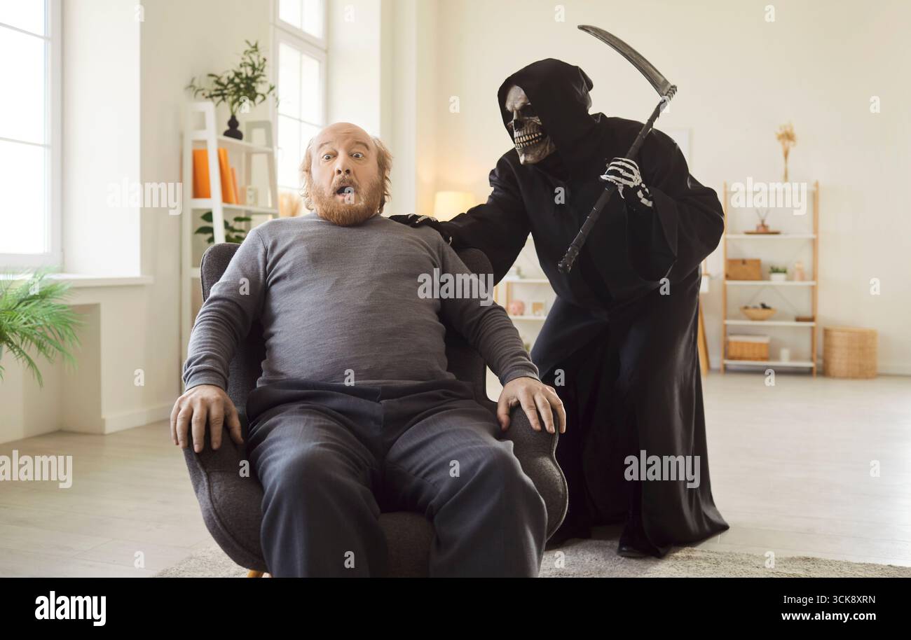 Uomo anziano spaventato seduto su una poltrona come un mietitore con un mantello nero che gli mette la mano sulla spalla. Foto Stock