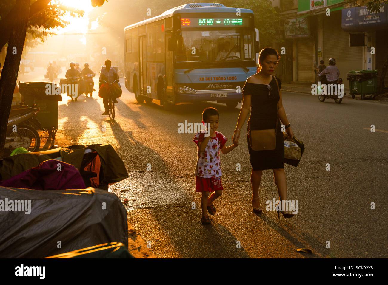 Madre che tiene in mano il bambino che attraversa la strada soleggiata di Hanoi, Vietnam, con autobus pubblici e motociclisti sullo sfondo. Hanoi, vietnam Foto Stock
