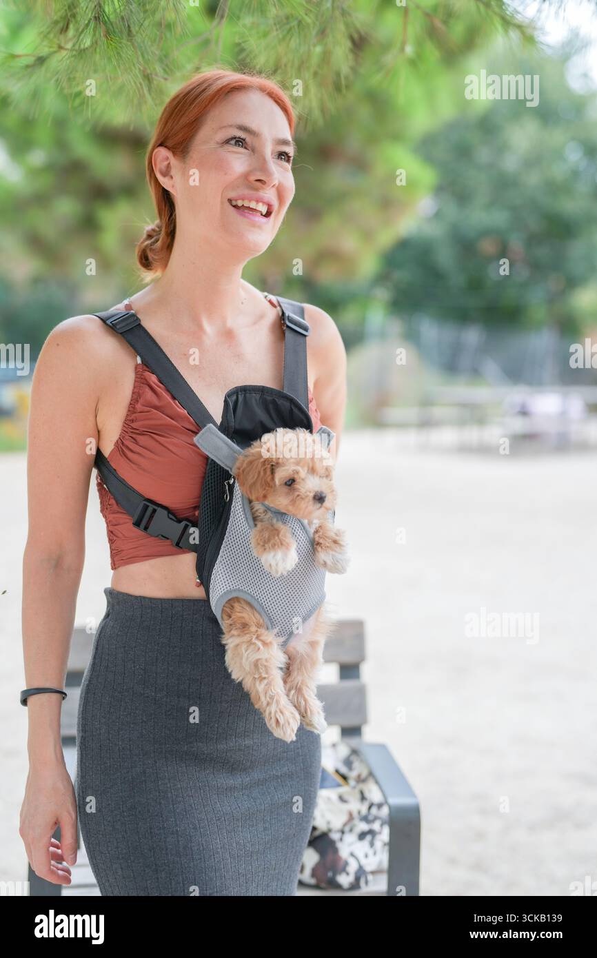 Una donna sorride mentre tiene un cane piccolo in un portabagagli. Il cane indossa un maglione e si sente a suo agio. La scena suggerisce un felice e rilassato Foto Stock