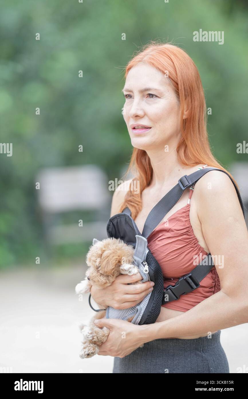 Una donna sta tenendo un cane piccolo in un portatore. Indossa una canotta rosa e pantaloni neri Foto Stock Una donna sta tenendo un cane piccolo in un portatore. Indossa una canotta rosa e pantaloni neri Foto Stock