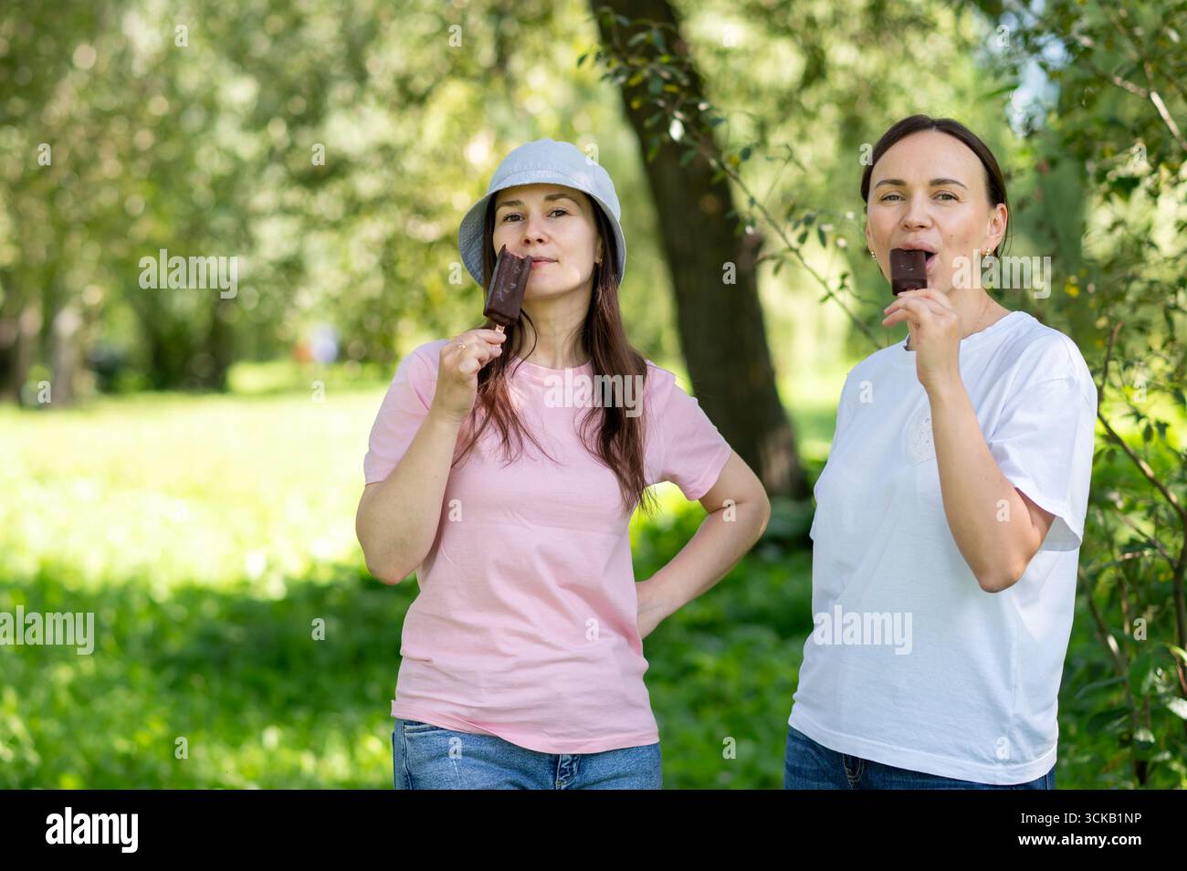 Due amiche si godono un gelato al cioccolato su bastoncini nel soleggiato parco estivo, un concetto di amicizia, svago e dolce dolce dolce rinfrescante Foto Stock