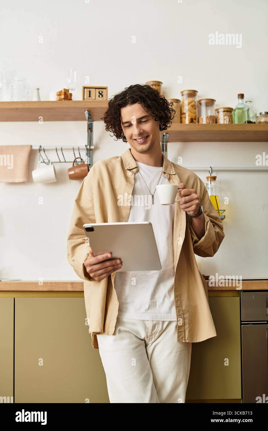 Un giovane affascinante in un abbigliamento casual sorride mentre tiene in mano una tazza e guarda il suo tablet. Foto Stock