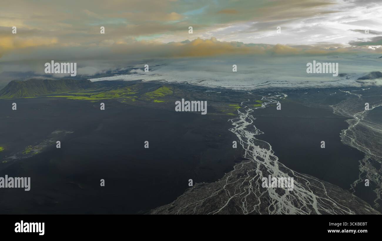 Prospettiva aerea del paesaggio vulcanico islandese con fiumi intrecciati, pianure scure, macchie di muschio verde e un ghiacciaio sotto basse nuvole. Foto Stock