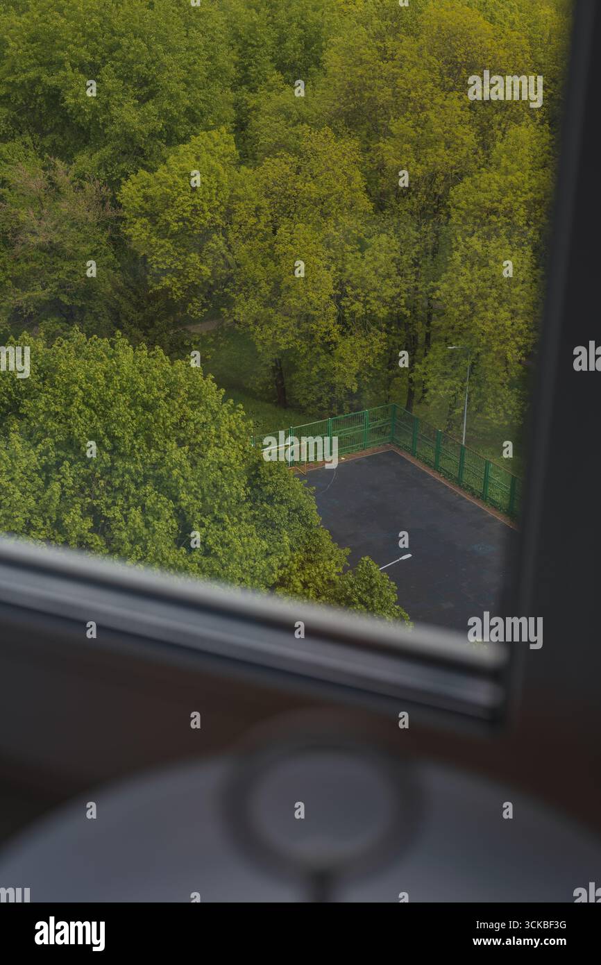Vista sopraelevata attraverso una finestra che mostra fitti alberi verdi e una piccola area asfaltata recintata, possibilmente un parcheggio o uno spazio ricreativo, sotto. Foto Stock