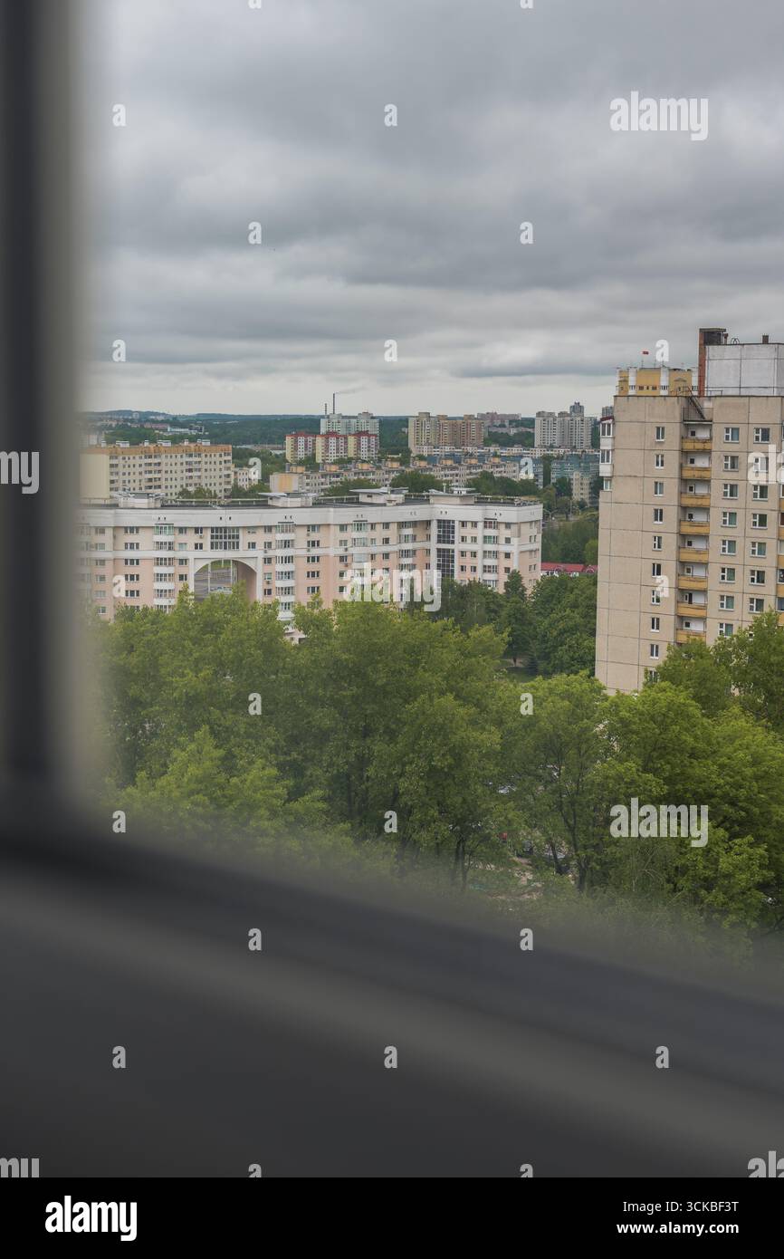 Vista urbana di Minsk, Bielorussia, che mostra moderni edifici residenziali con facciate chiare, incorniciati da alberi verdi e un cielo coperto attraverso una finestra. Foto Stock