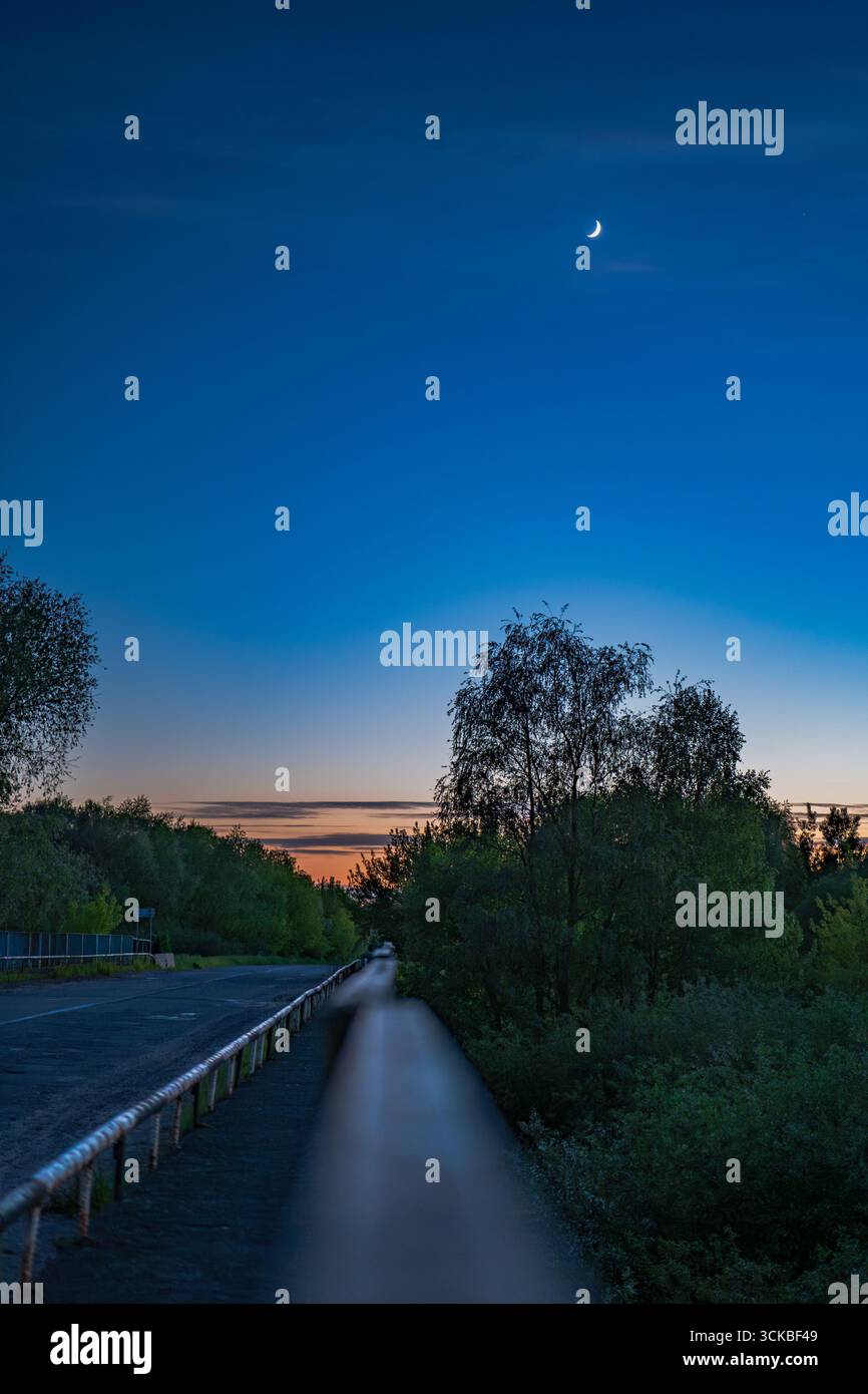 Una tranquilla strada rurale in Bielorussia al crepuscolo, delimitata da alberi e ringhiera di metallo. Una luna crescente brilla nel cielo blu profondo sopra un orizzonte luminoso Foto Stock