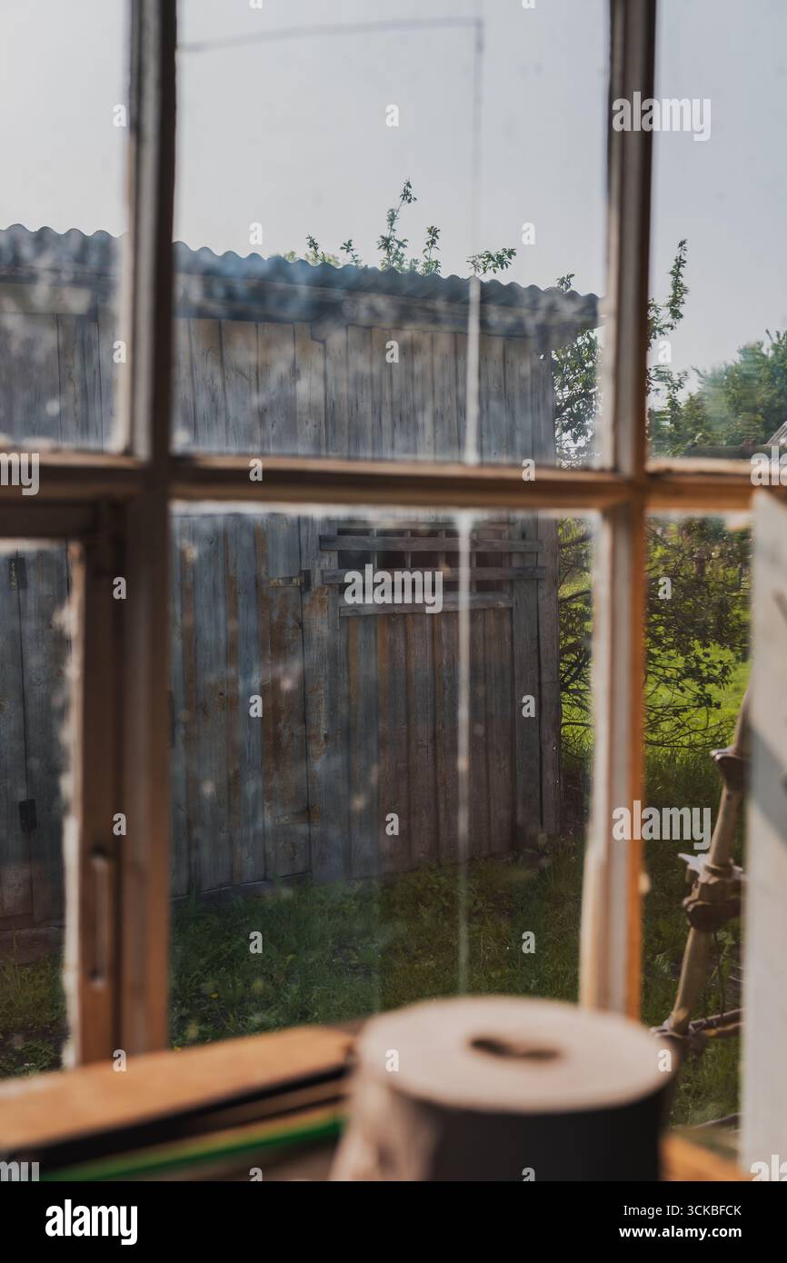 Una finestra incorniciata in legno rivela un capannone con tetto metallico, circondato da erba e alberi. Un rotolo di carta poggia sul davanzale interno. Foto Stock