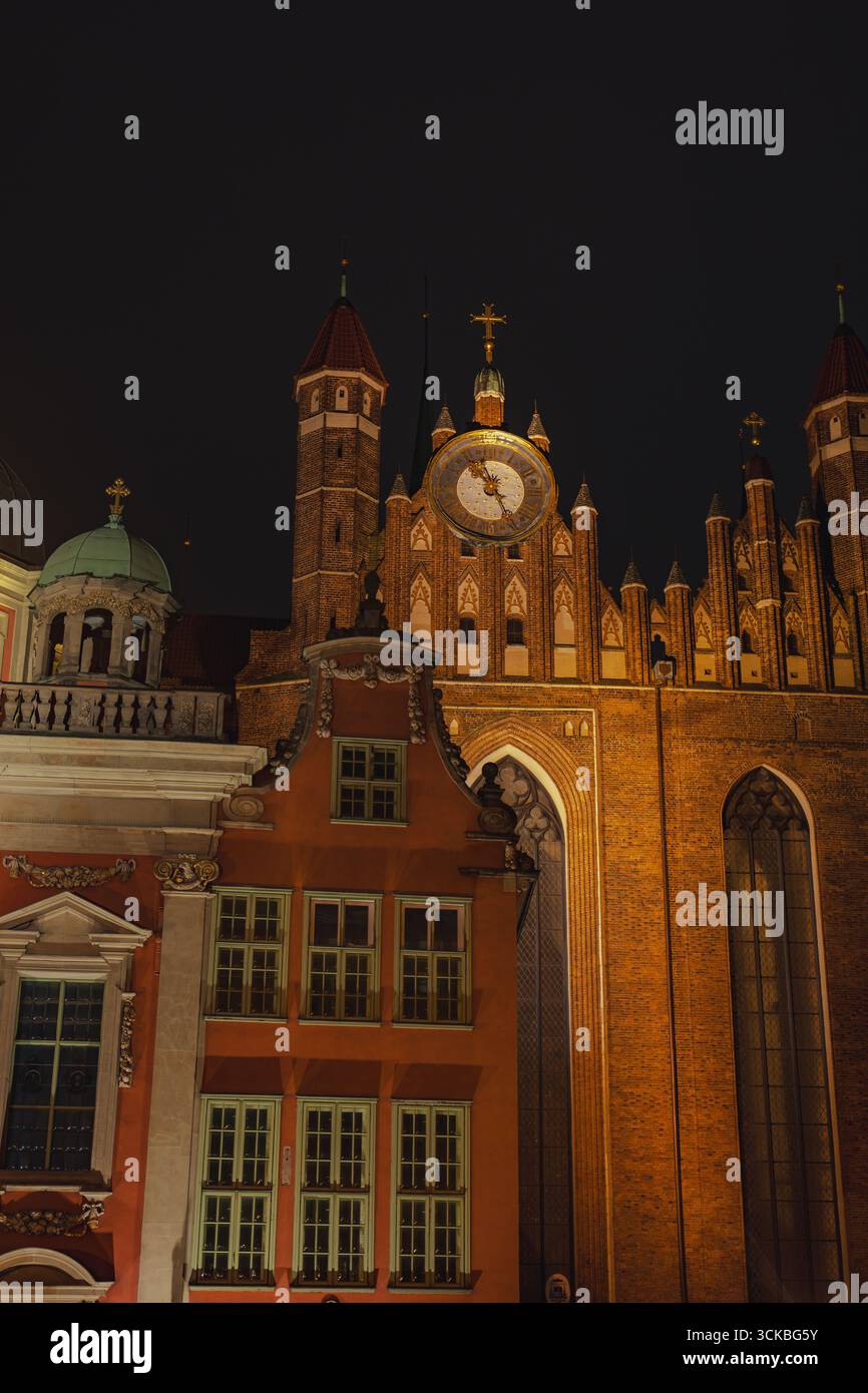 Municipio principale illuminato con torre dell'orologio a Danzica, Polonia, circondato da dettagli gotici, un edificio rosso e una struttura a cupola sotto un cielo scuro. Foto Stock