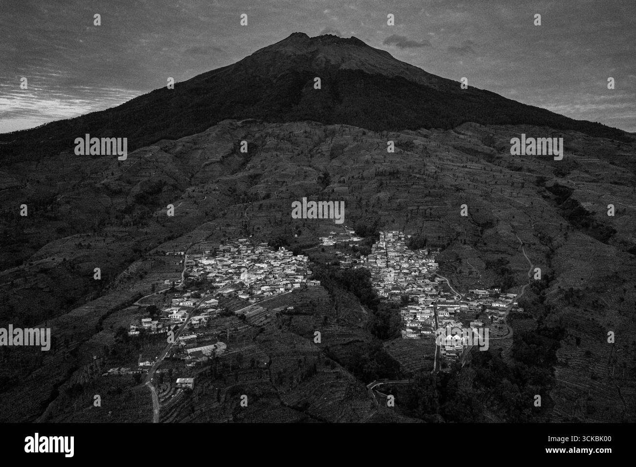 Vista aerea di un villaggio annidato in un terreno montuoso con un prominente vulcano sullo sfondo, catturato in bianco e nero Foto Stock