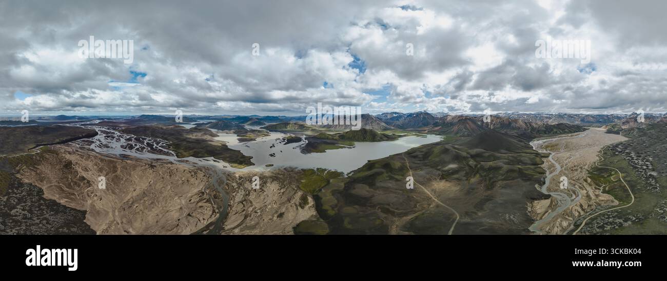 I fiumi intrecciati scorrono attraverso il terreno vulcanico con muschio verde, un lago centrale, montagne innevate e colline riolite sotto un cielo nuvoloso. Foto Stock