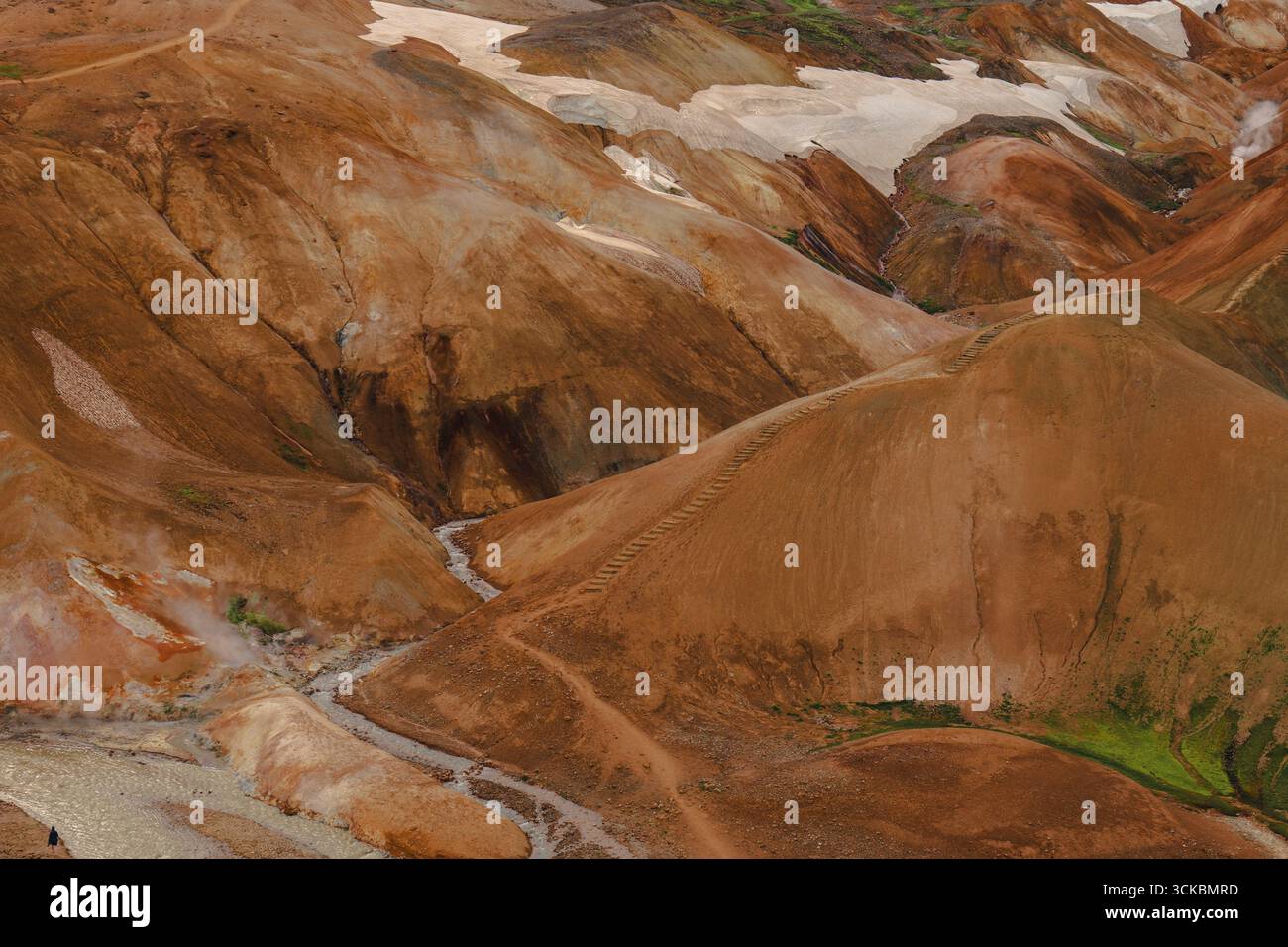 Vivaci colline di riolite arancioni, rosse e marroni nel Kerlingarfjoll islandese, con un torrente tortuoso, vapore geotermico, chiazze di neve e un sentiero escursionistico. Foto Stock