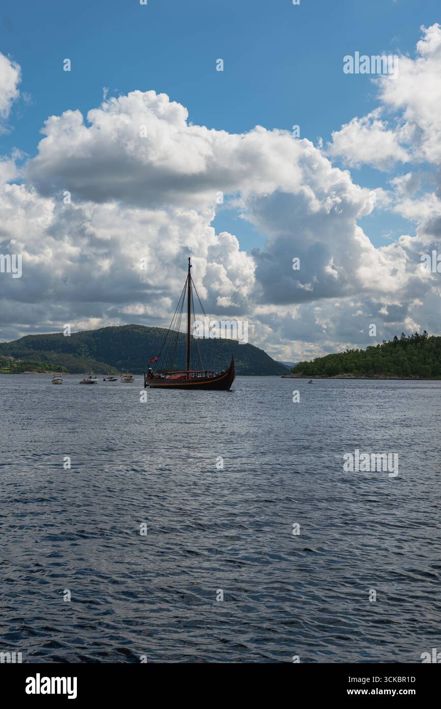 Una nave vichinga con una prua curva e un solo albero galleggia su acque calme, circondata da barche più piccole, con verdi colline e un cielo parzialmente nuvoloso nel bac Foto Stock