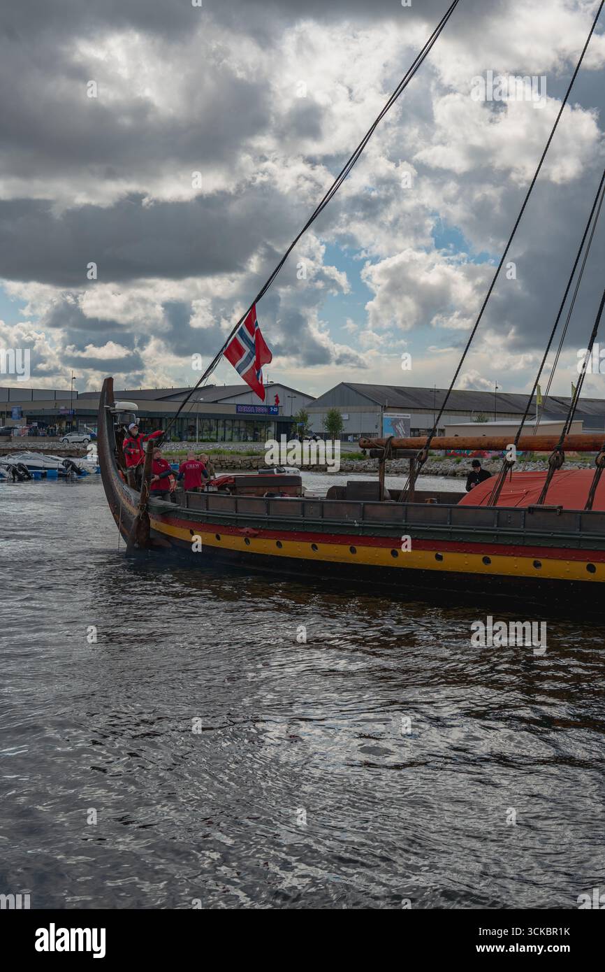 Una nave vichinga con prua in legno curva e scafo giallo rosso naviga vicino a un moderno lungomare. Le persone in rosso sono a bordo sotto un cielo parzialmente nuvoloso. Foto Stock