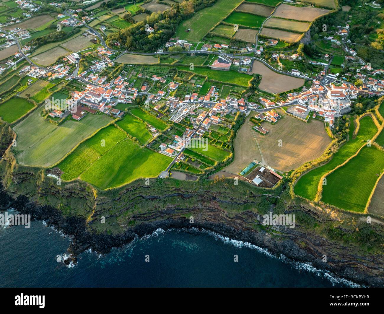 Vista aerea del villaggio annidato tra vivaci campi verdi e la costa scura e frastagliata, Feiteras, Azzorre, Portogallo. Foto Stock