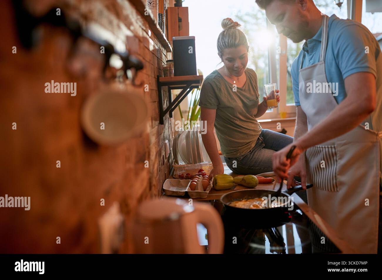 Marito che prepara la colazione in una padella, uova strapazzate, mentre la moglie è seduta sul bancone a guardare cosa sta facendo. Insieme, stile di vita, casa Foto Stock