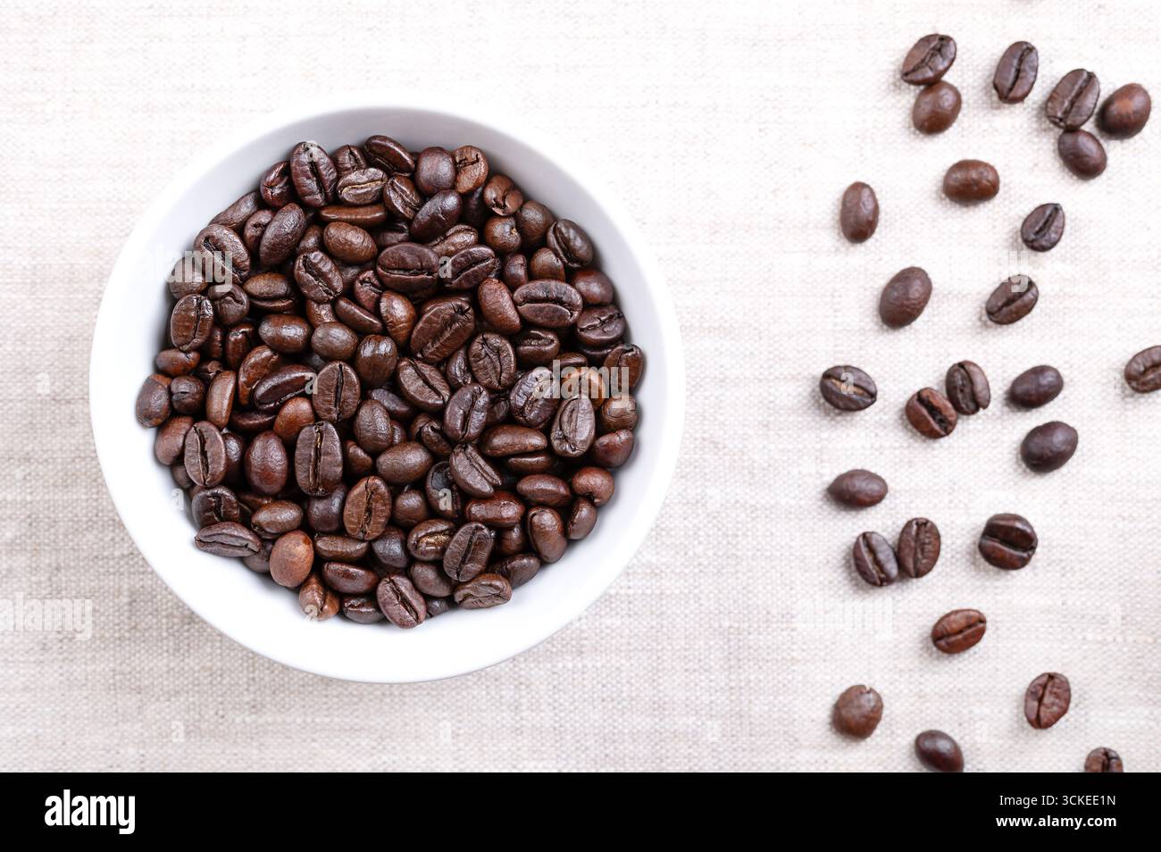 Chicchi di caffè arabica tostati, in un recipiente bianco, su lino. Semi interi di frutti di bosco tostati di Coffea arabica. Foto Stock