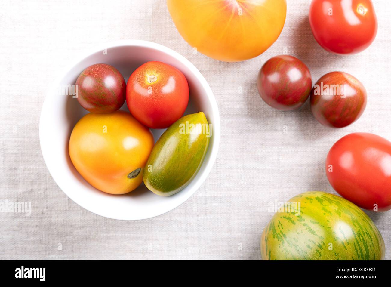 Pomodori freschi e maturi di varie varietà, colori e dimensioni, in una ciotola bianca, su tessuto di lino. Foto Stock