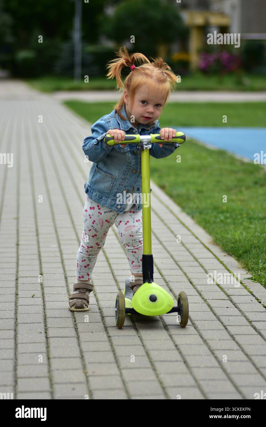 Bambina dai capelli rossi con giacca in denim e leggings in sella a uno scooter verde sul marciapiede lastricato. Bambino che pratica l'equilibrio e la coordinazione mentre si diverte Foto Stock