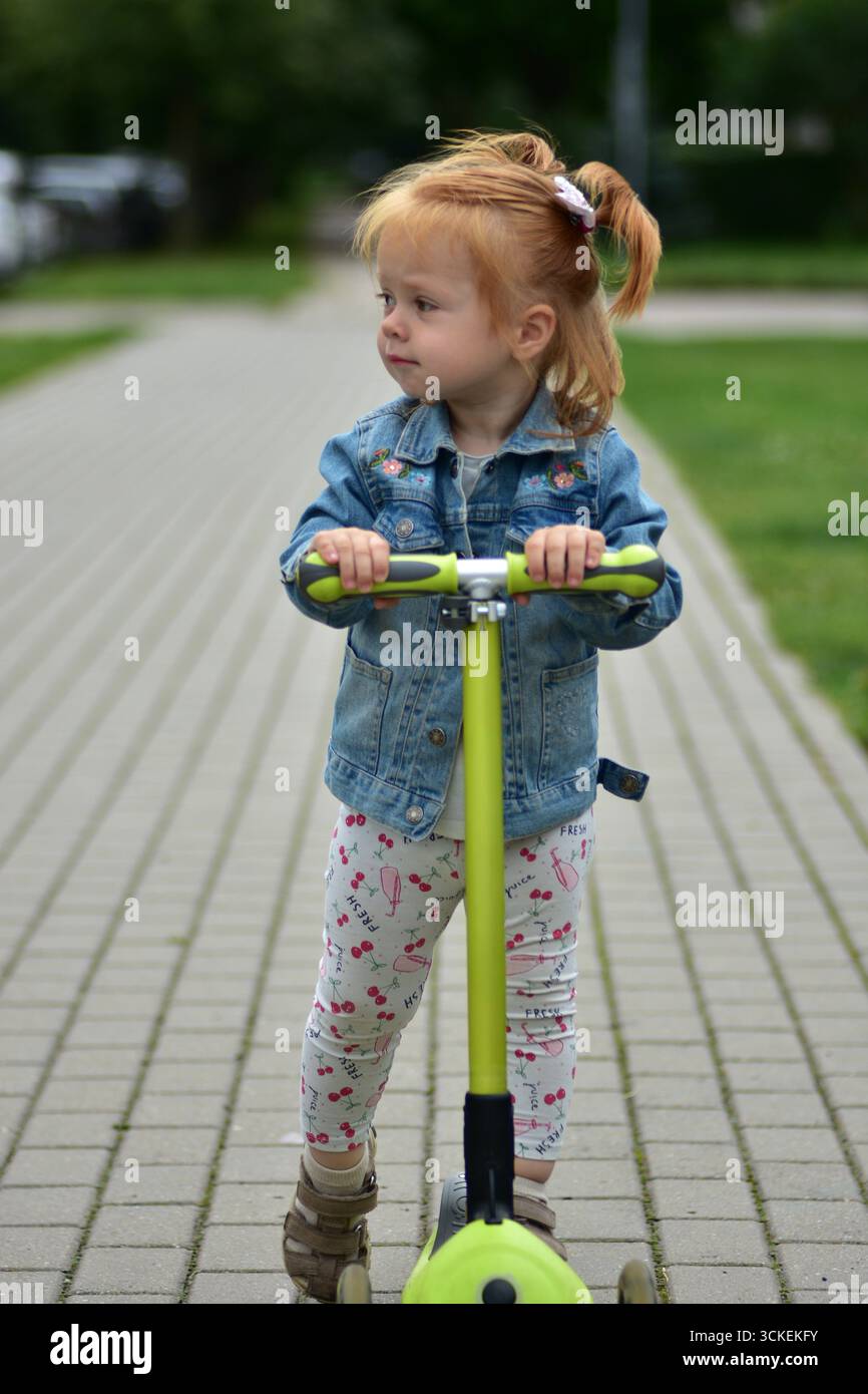 Bambina dai capelli rossi con giacca in denim e leggings in sella a uno scooter verde sul marciapiede lastricato. Bambino che pratica l'equilibrio e la coordinazione mentre si diverte Foto Stock