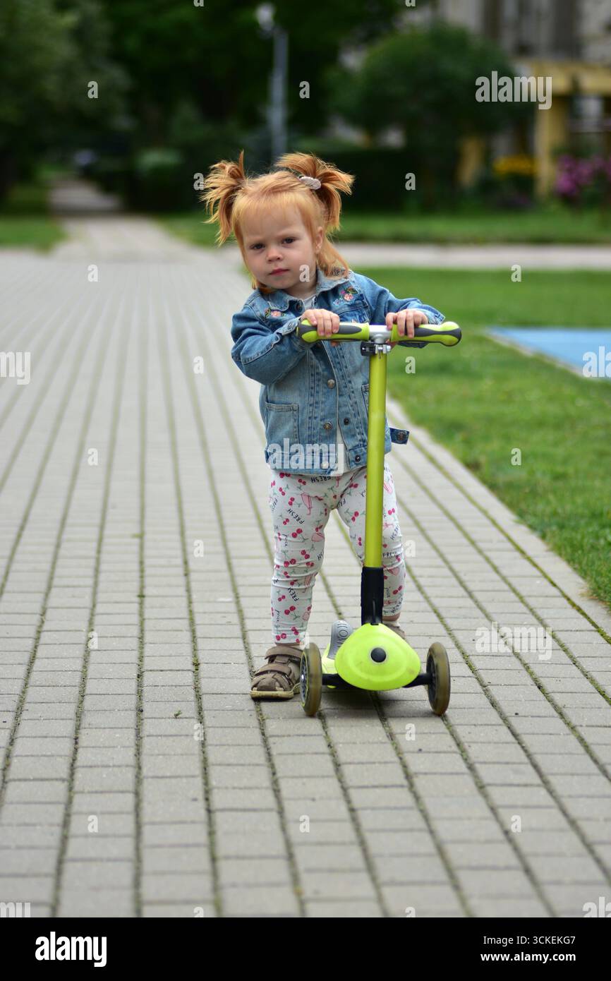 Bambina dai capelli rossi con giacca in denim e leggings in sella a uno scooter verde sul marciapiede lastricato. Bambino che pratica l'equilibrio e la coordinazione mentre si diverte Foto Stock
