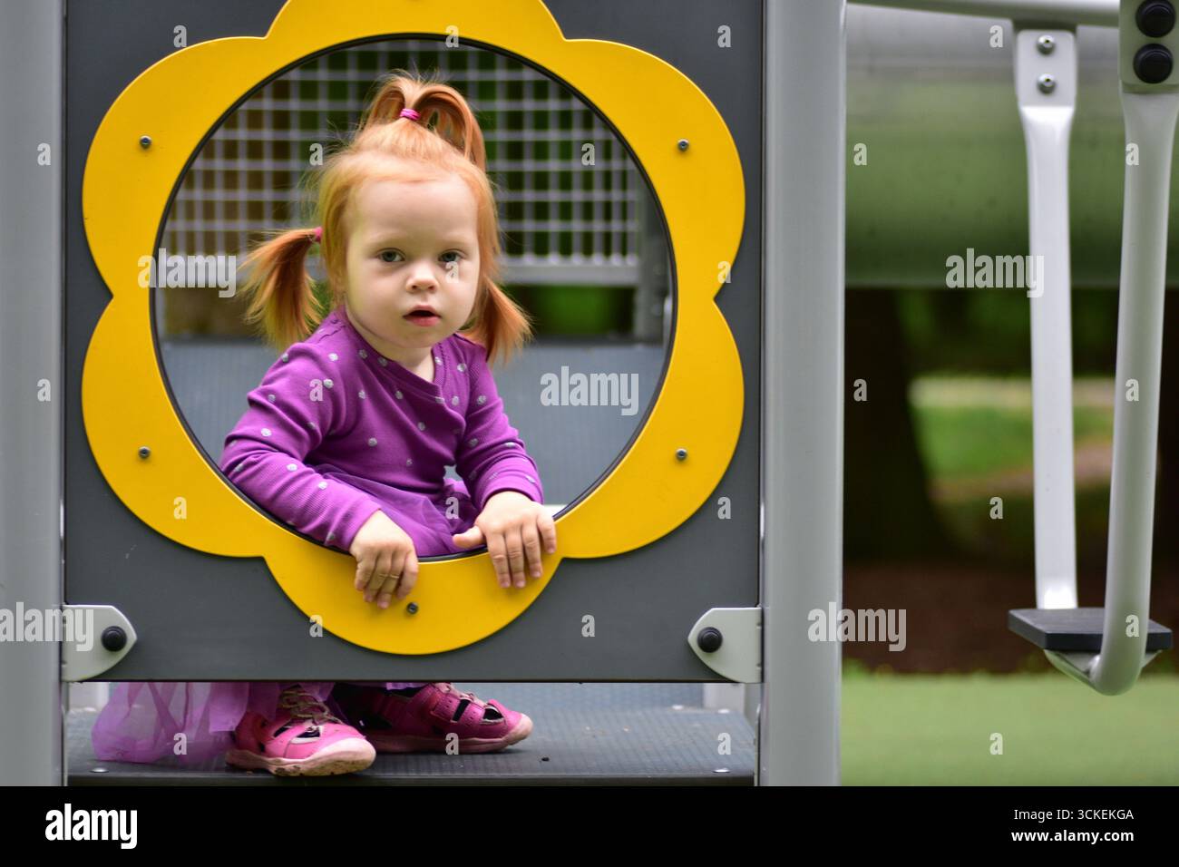 Bambina dai capelli rossi in abito viola seduta all'interno della cornice circolare gialla del parco giochi. Un bambino curioso mentre si diverte a giocare all'aperto in estate Foto Stock