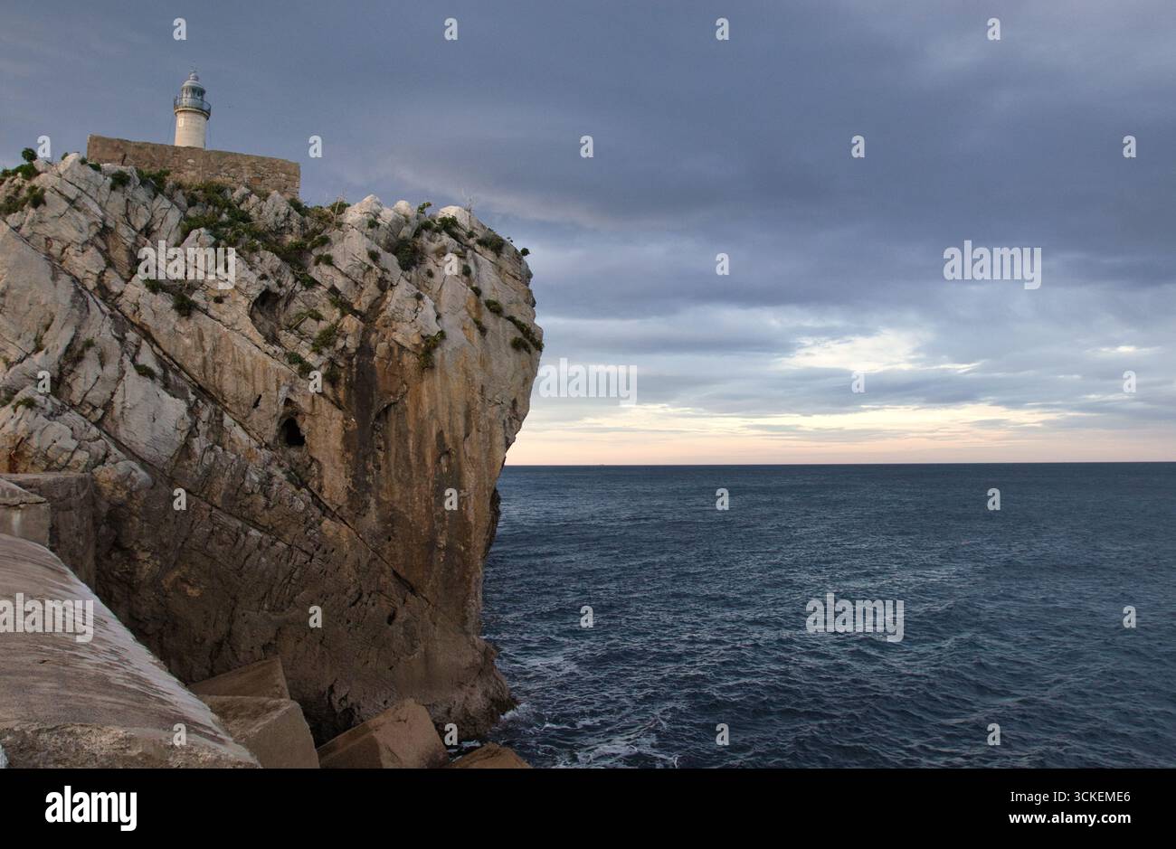 Faro remoto su una spettacolare scogliera sul mare - Un piccolo faro bianco sorge su una scogliera rocciosa aspra che si affaccia su un mare scuro e ondulato sotto un suggestivo clo Foto Stock