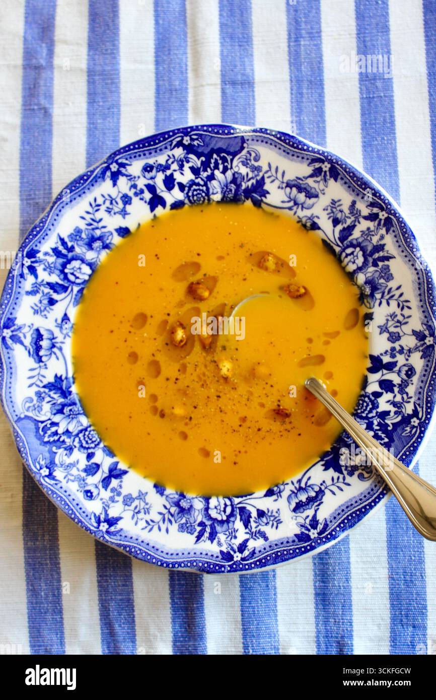Ciotola blu e bianca in porcellana di zuppa di zucca guarnita con semi di zucca arrostiti e olio d'oliva, su una tovaglia di lino a righe blu e bianche. Foto Stock