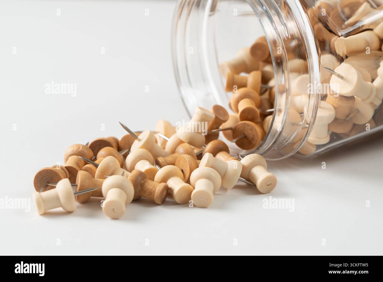 Un vaso in vetro trasparente è rovesciato su un lato, con una collezione di spinotti in legno che fuoriescono su una superficie bianca. I perni sono un mix di toni di legno chiari e medi, ciascuno con una punta metallica argentata. Foto Stock