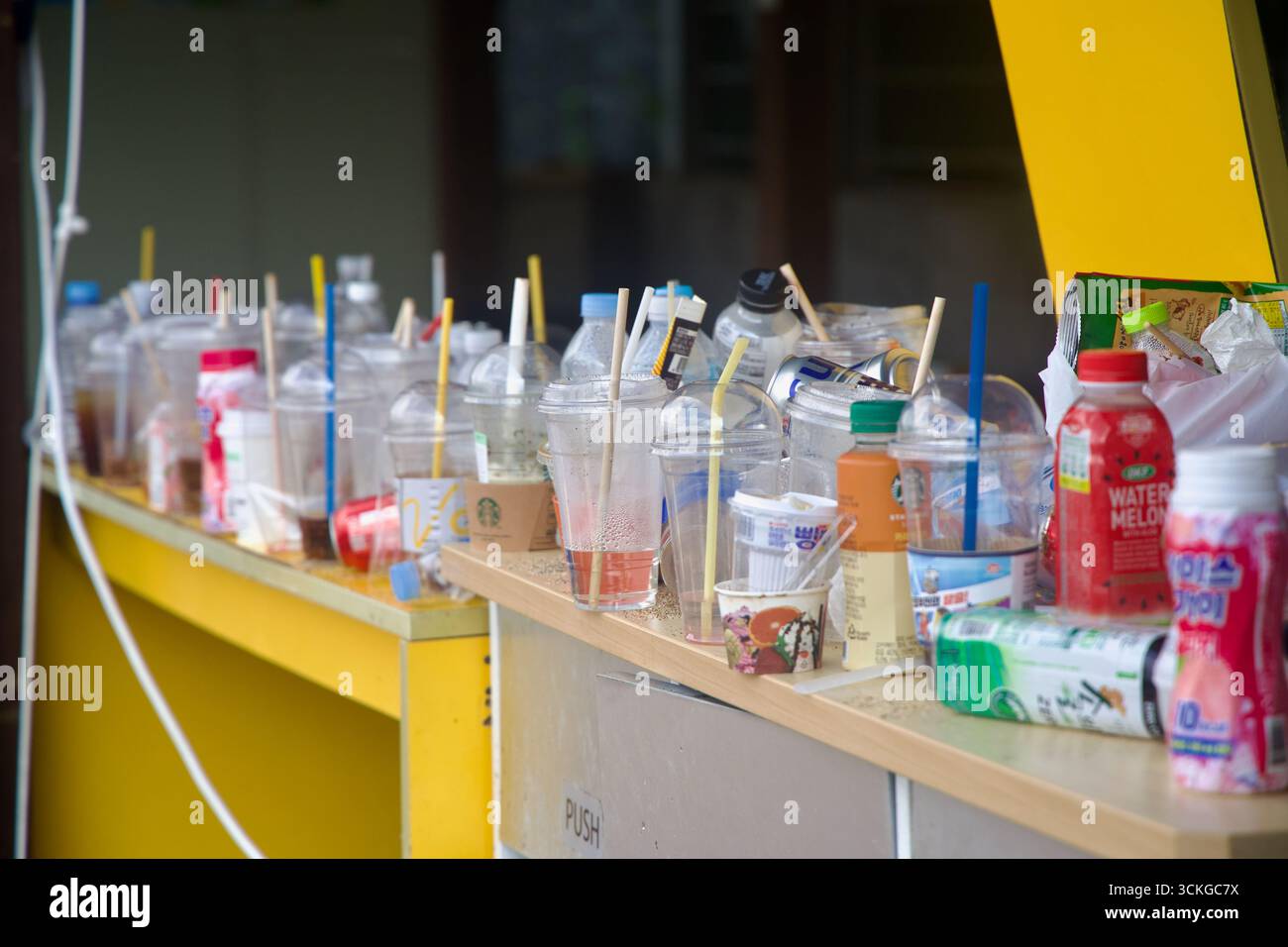 Una fila ingombrante di contenitori per bevande scartate, tra cui bicchieri, bottiglie e cartoni di plastica con marchi visibili, allinea un bancone giallo all'aperto Foto Stock