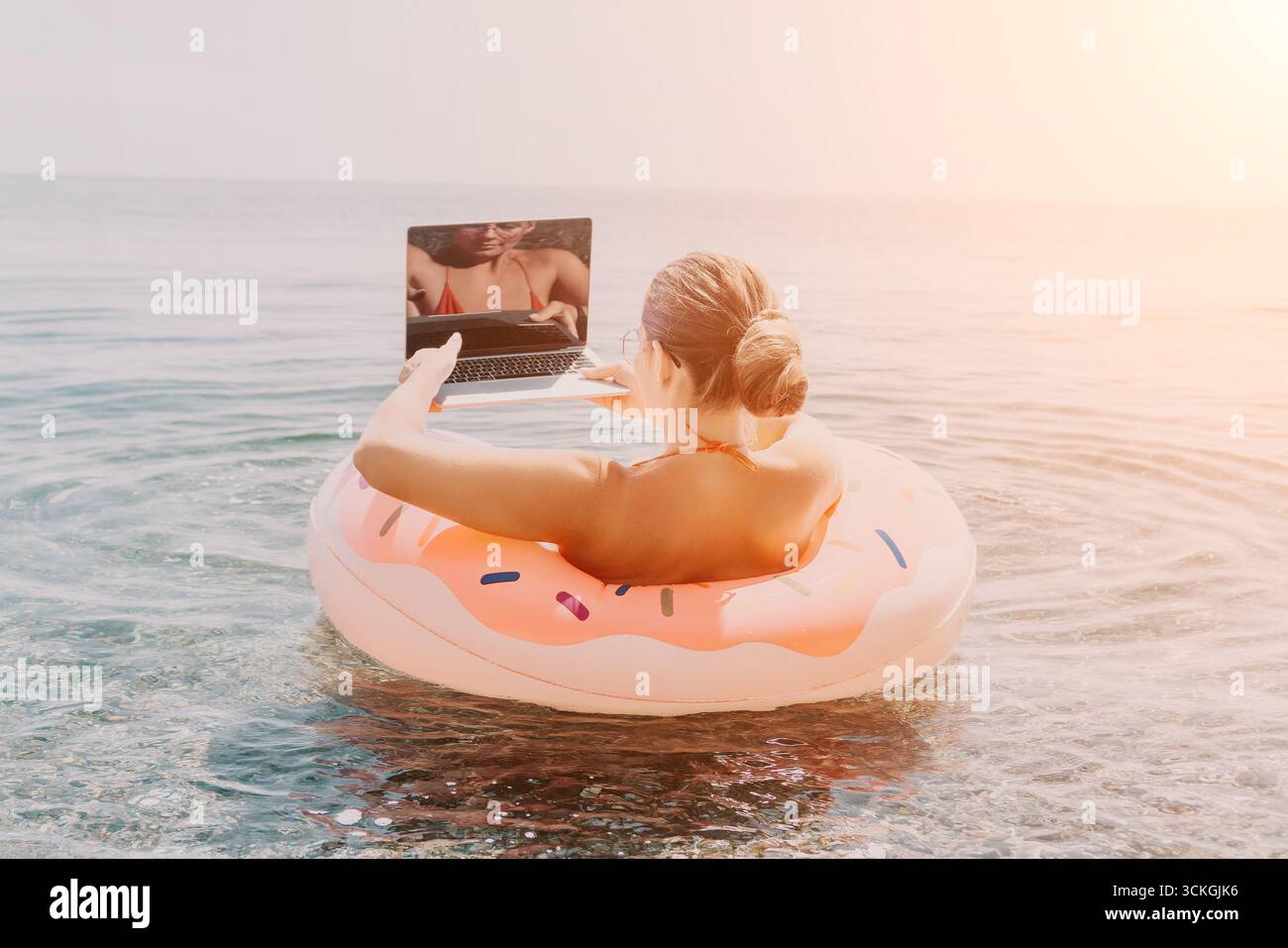 Woman laptop Beach - lavora in remoto su un laptop mentre galleggia in ciambella gonfiabile nell'oceano al tramonto. Foto Stock