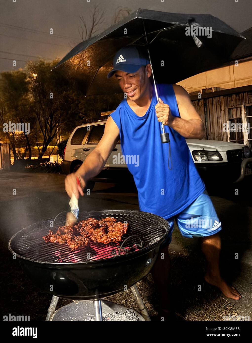 Uomo sorridente che cucina al barbecue all'aperto sotto la pioggia con ombrello, Cairns, Queensland, Australia. No MR o PR Foto Stock