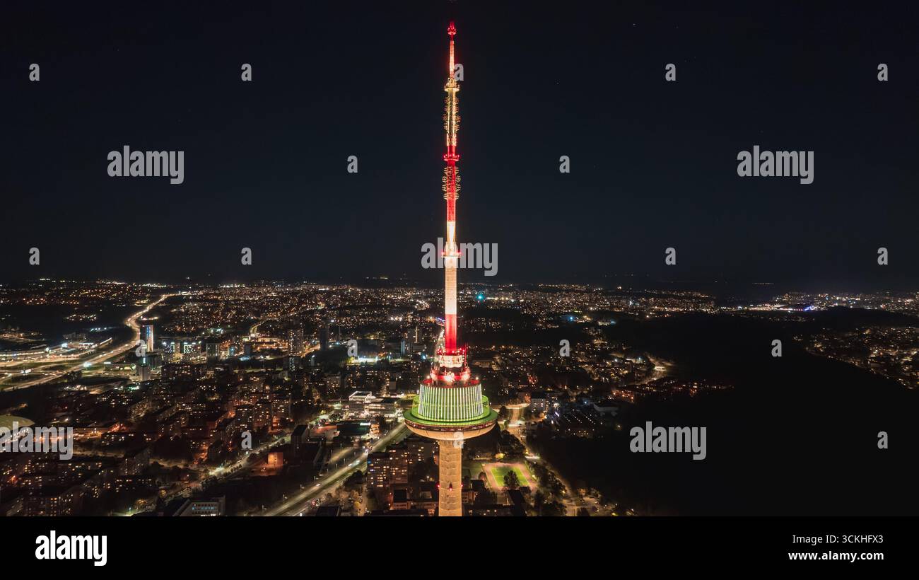La torre della televisione di Vilnius si erge illuminata con luci rosse e verdi contro un cielo scuro, circondata dalle luci urbane e dalle strade della città di notte. Foto Stock