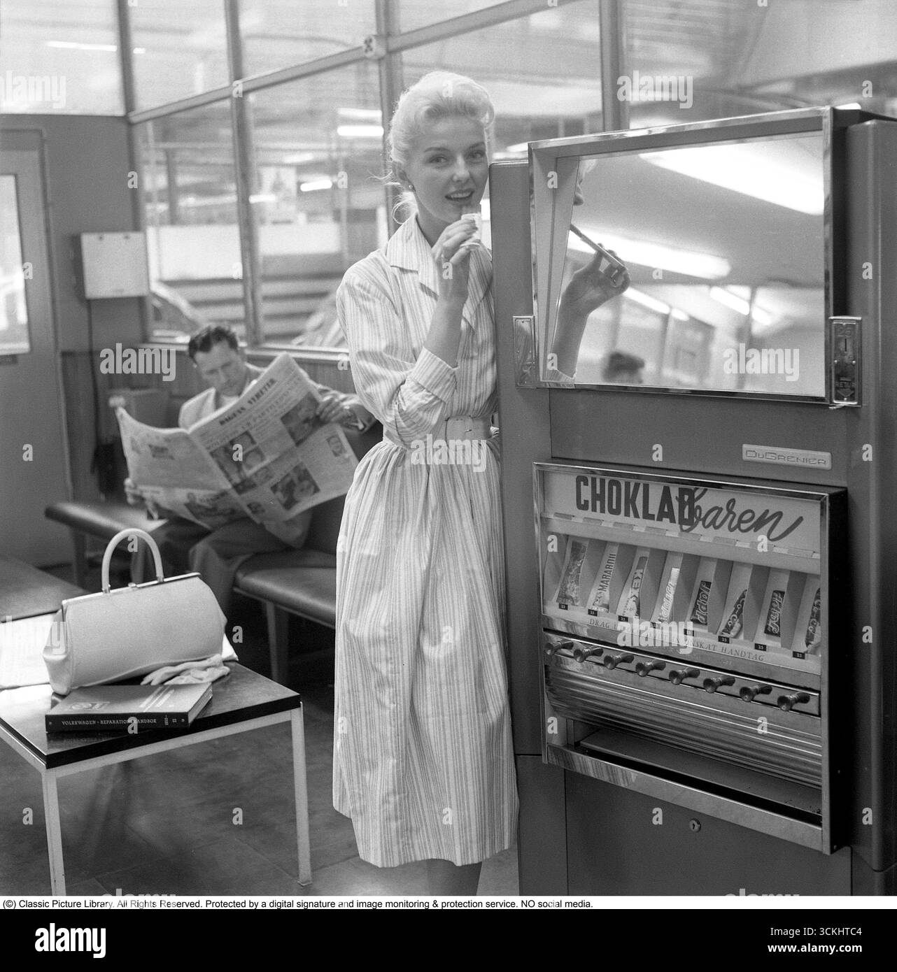 Distributore automatico 1957. Una donna bionda si appoggia al distributore automatico che offre barrette di cioccolato di diversi tipi. La tipica gonna completa popolare per le donne nel 1957 era una gonna larga, svasata A-line o circolare, spesso fissata in vita con una cintura per creare una silhouette a clessidra. Realizzato in tessuti come cotone, tulle o chiffon, in genere è caduto a metà polpaccio ed è stato indossato con petticoats per un maggiore volume. Lo stile era vivace, femminile e iconico della moda "New Look" degli anni '1950. Conard rif. 3496 Foto Stock