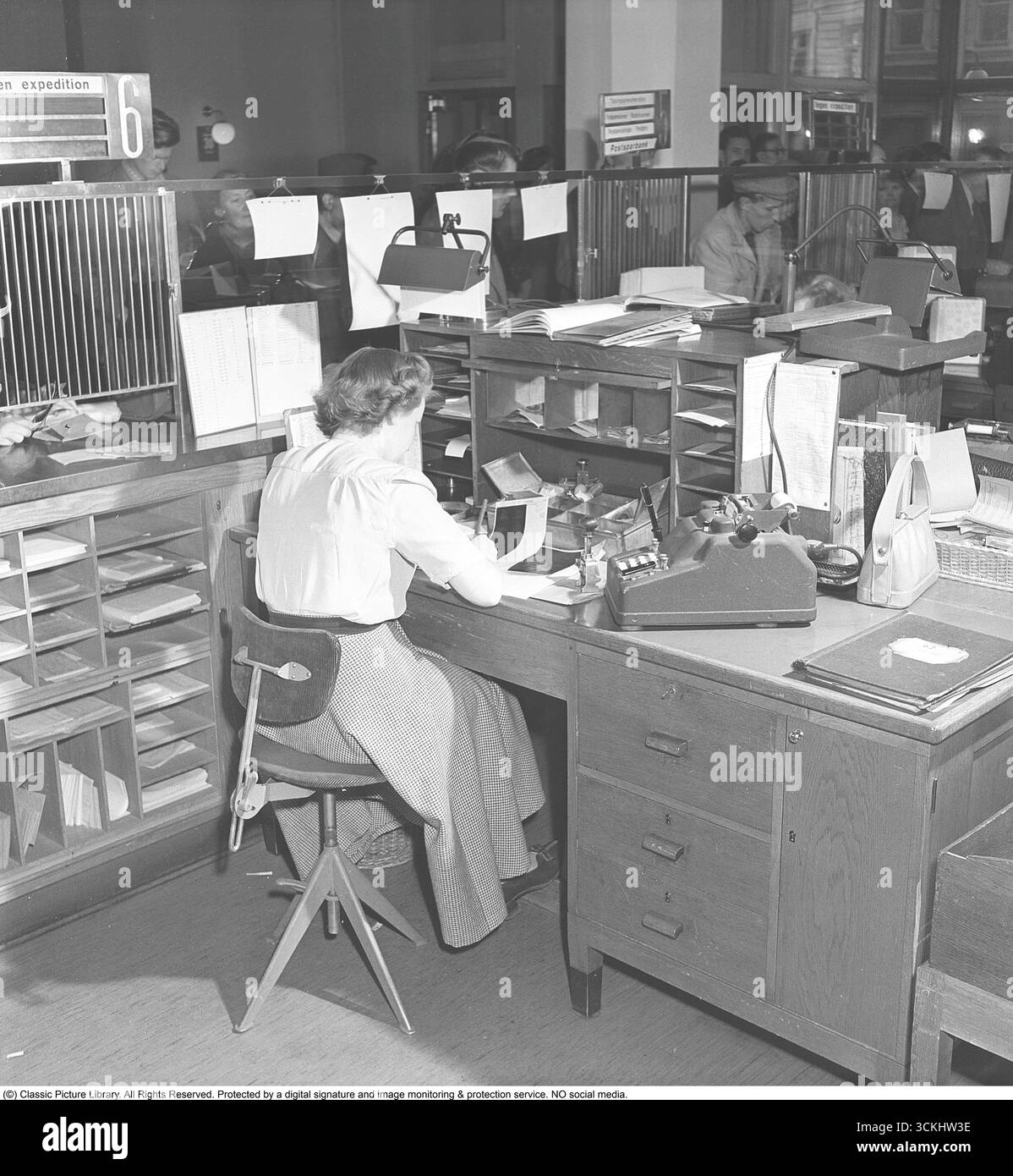 Interno di una banca con una giovane donna dietro il bancone dell'ufficio della banca. Si vede aiutare un cliente che si vede in piedi alla cassa tenendo in mano il suo libretto di risparmio. Un libretto di risparmio è un opuscolo fisico utilizzato dai singoli individui per registrare le transazioni in un conto di risparmio, comune nelle banche e nella vita quotidiana prima del digital banking. Simboleggia la storia finanziaria e la cultura del risparmio personale, evocando nostalgia per le pratiche bancarie tradizionali. 1949. Kristoffersson rif. AT21-1 Foto Stock