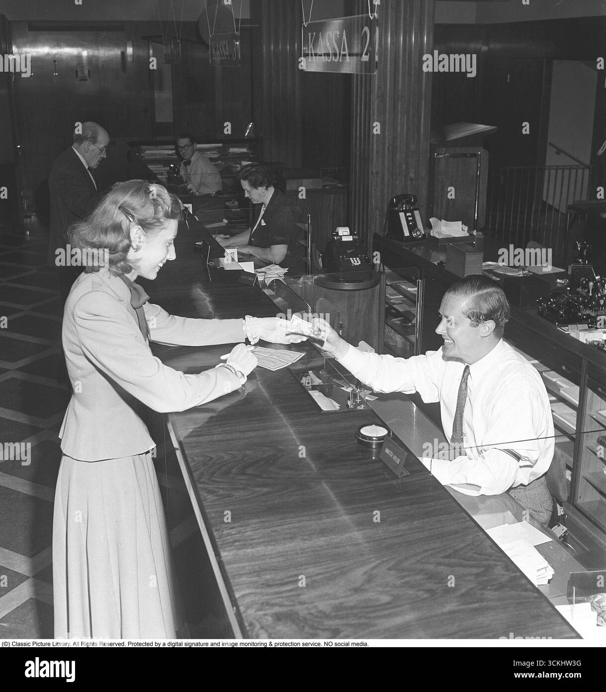 L'interno di una banca con un uomo dietro il bancone nell'ufficio della banca. Viene visto aiutare un cliente che viene visto in piedi alla cassa. L'uso e la gestione delle banconote fisiche erano comuni nelle banche e nella vita quotidiana prima del digital banking. Simboleggia la storia finanziaria e la cultura del risparmio personale, evocando nostalgia per le pratiche bancarie tradizionali. 1949. Kristoffersson rif. AS17-3 Foto Stock