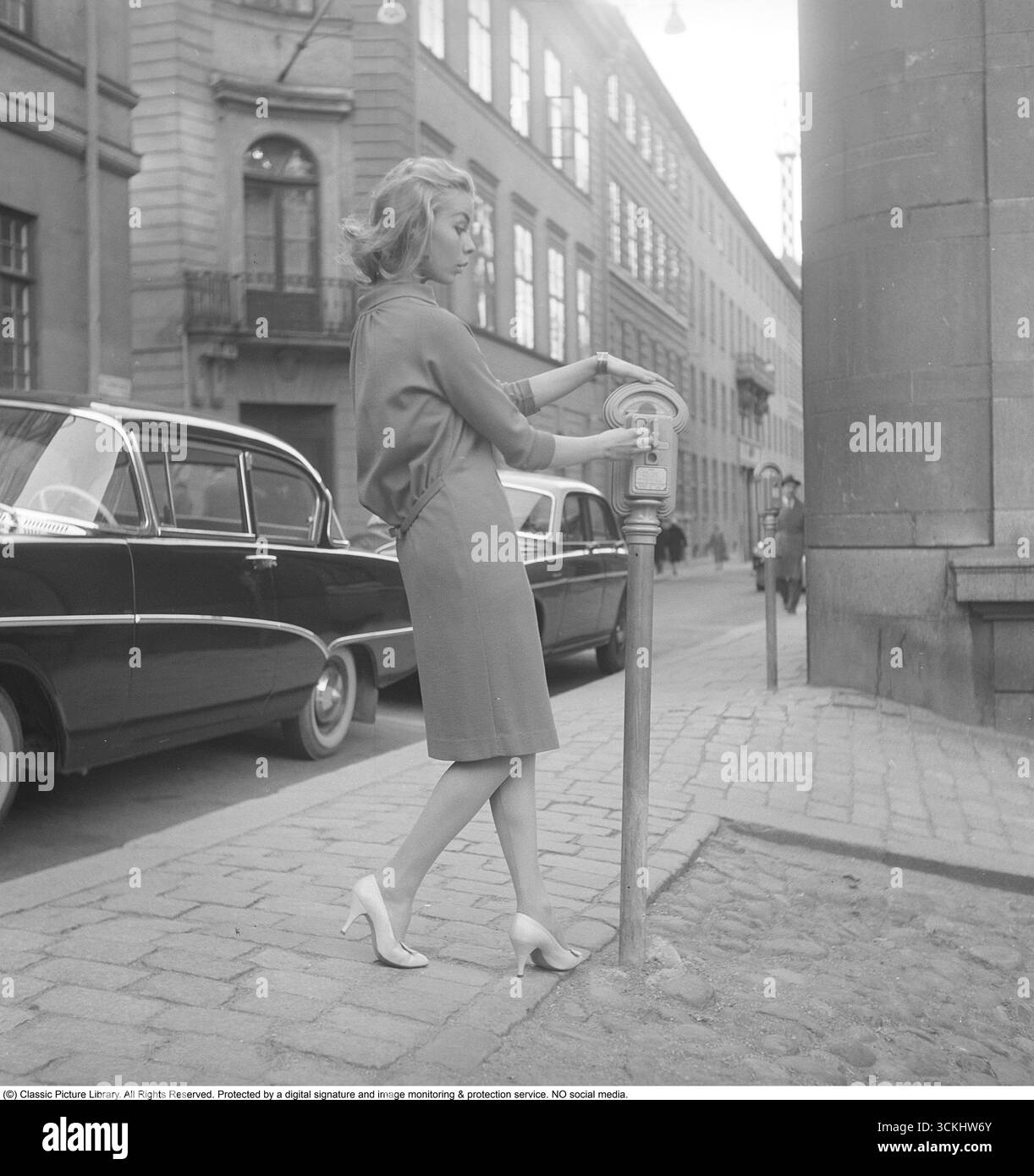 Nel 1956, quando i parchimetri richiedevano ancora monete, una giovane donna parcheggia la sua auto in una strada trafficata e inserisce le monete nel parcheggio accanto al suo posto auto. Dopo aver inserito le monete, gira il quadrante per impostare il tempo di parcheggio a pagamento e l'orologio del contatore inizia il conto alla rovescia. Questa scena cattura il fascino della vita urbana della metà del XX secolo, con auto d'epoca e contatori meccanici che riflettono un'era più semplice e basata sul denaro. Kristoffersson rif. BZ28-2. Foto Stock